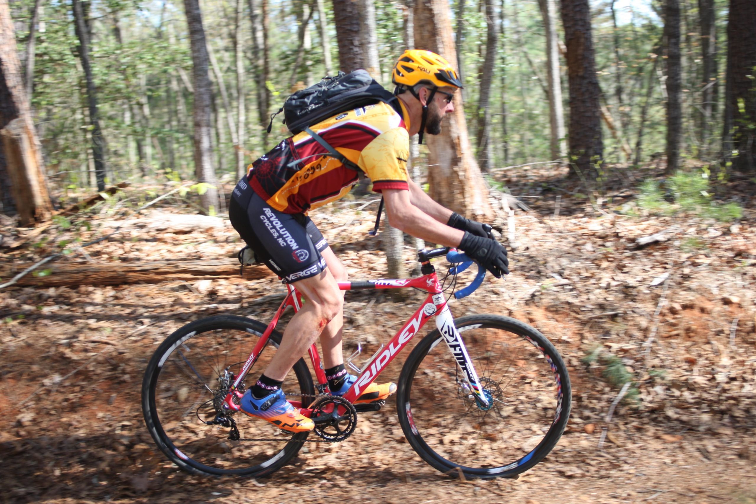 A cyclist riding a pink bike through a wooded trail. The cyclist is wearing a colorful jersey, shorts, gloves, and a helmet, and has a backpack. The background features trees and a dirt path, suggesting an outdoor mountain biking activity. Dark Mountain Trail mountain bike trail.