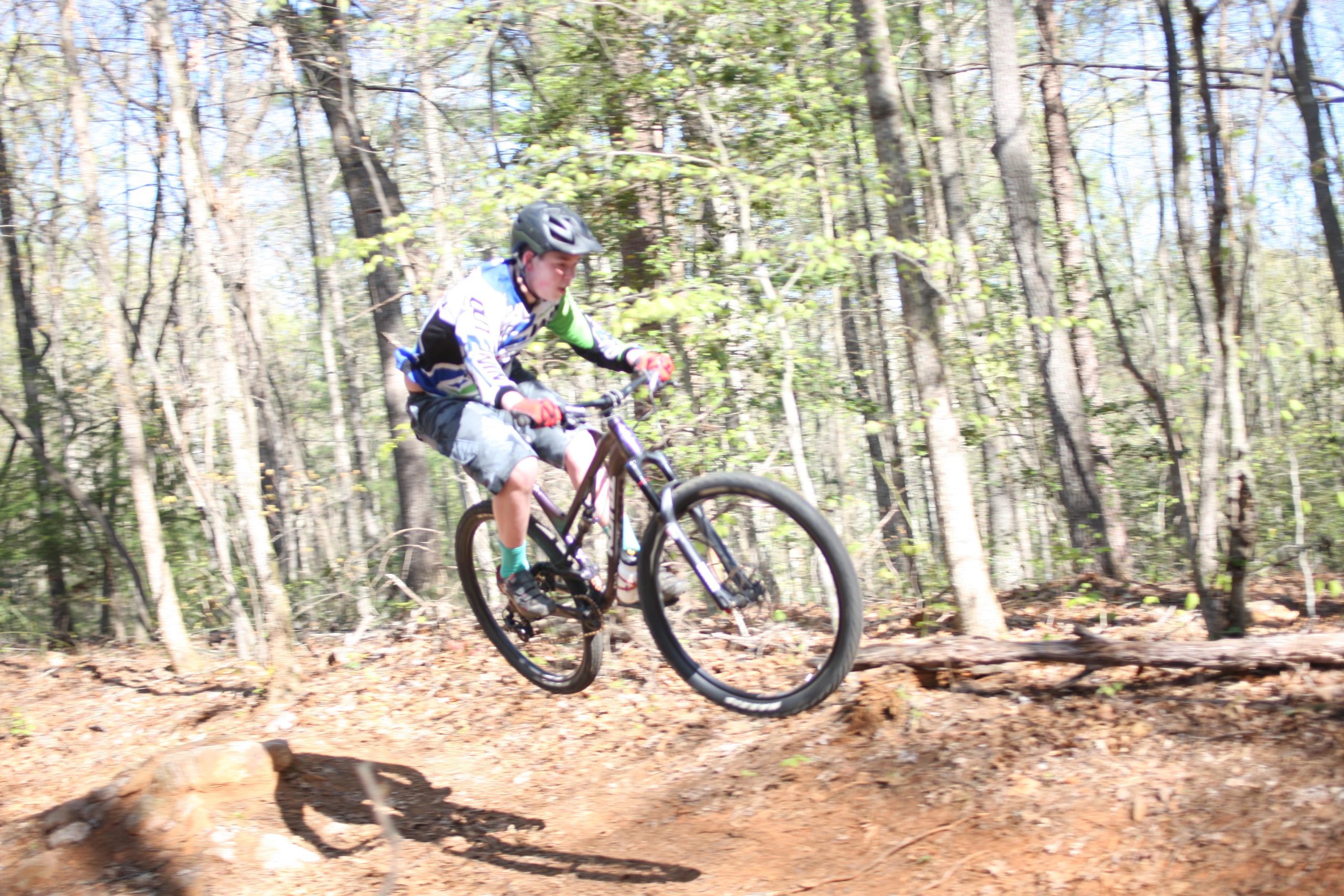 A young cyclist in a helmet performs a jump on a mountain bike, capturing the thrill of the sport. The scene is set in a lush forest with trees in the background, and the ground is covered with dirt and fallen leaves. The cyclist is wearing a blue and green jersey and shorts, showcasing an action shot filled with motion and excitement. Dark Mountain Trail mountain bike trail.