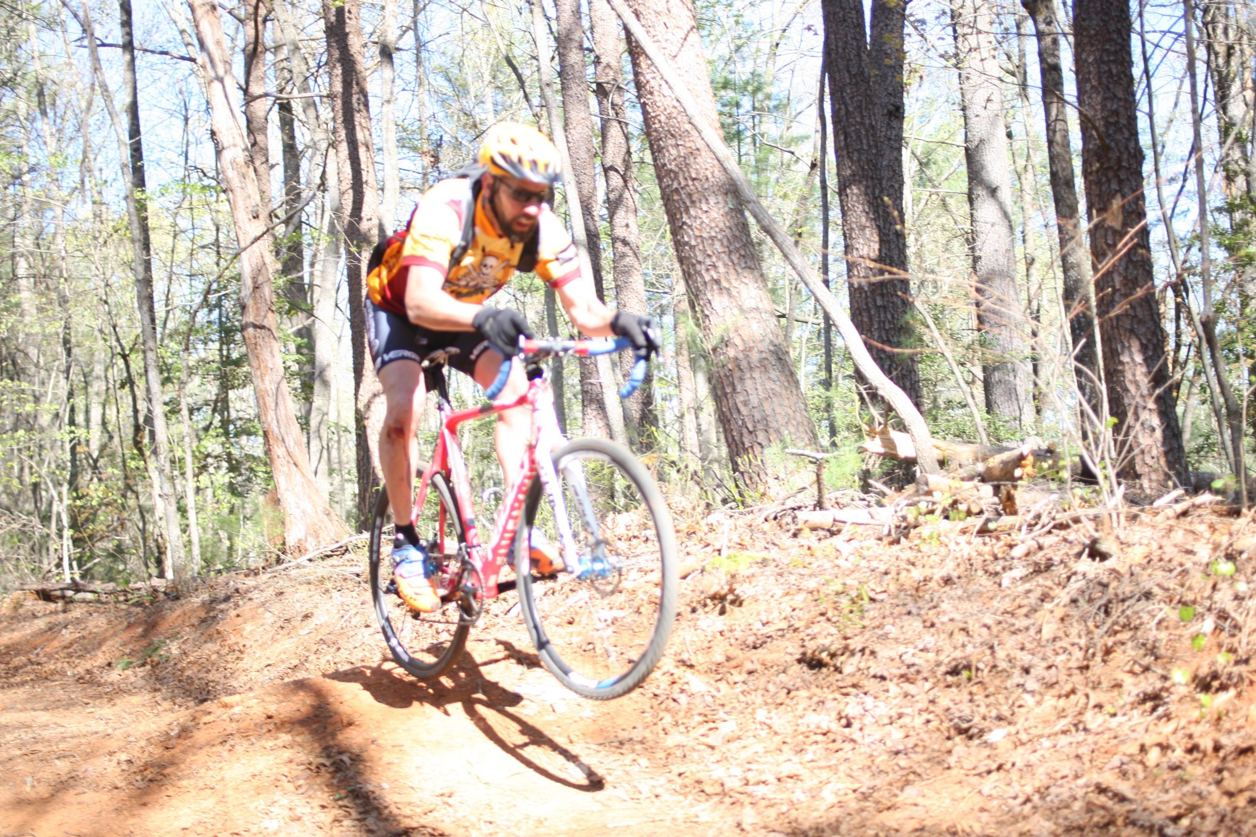 A cyclist in a yellow and red jersey soars over a dirt trail in a wooded area, navigating a bend with trees and sunlight filtering through the leaves. The cyclist is airborne, riding a pink bike and wearing a helmet and gloves. Dark Mountain Trail mountain bike trail.