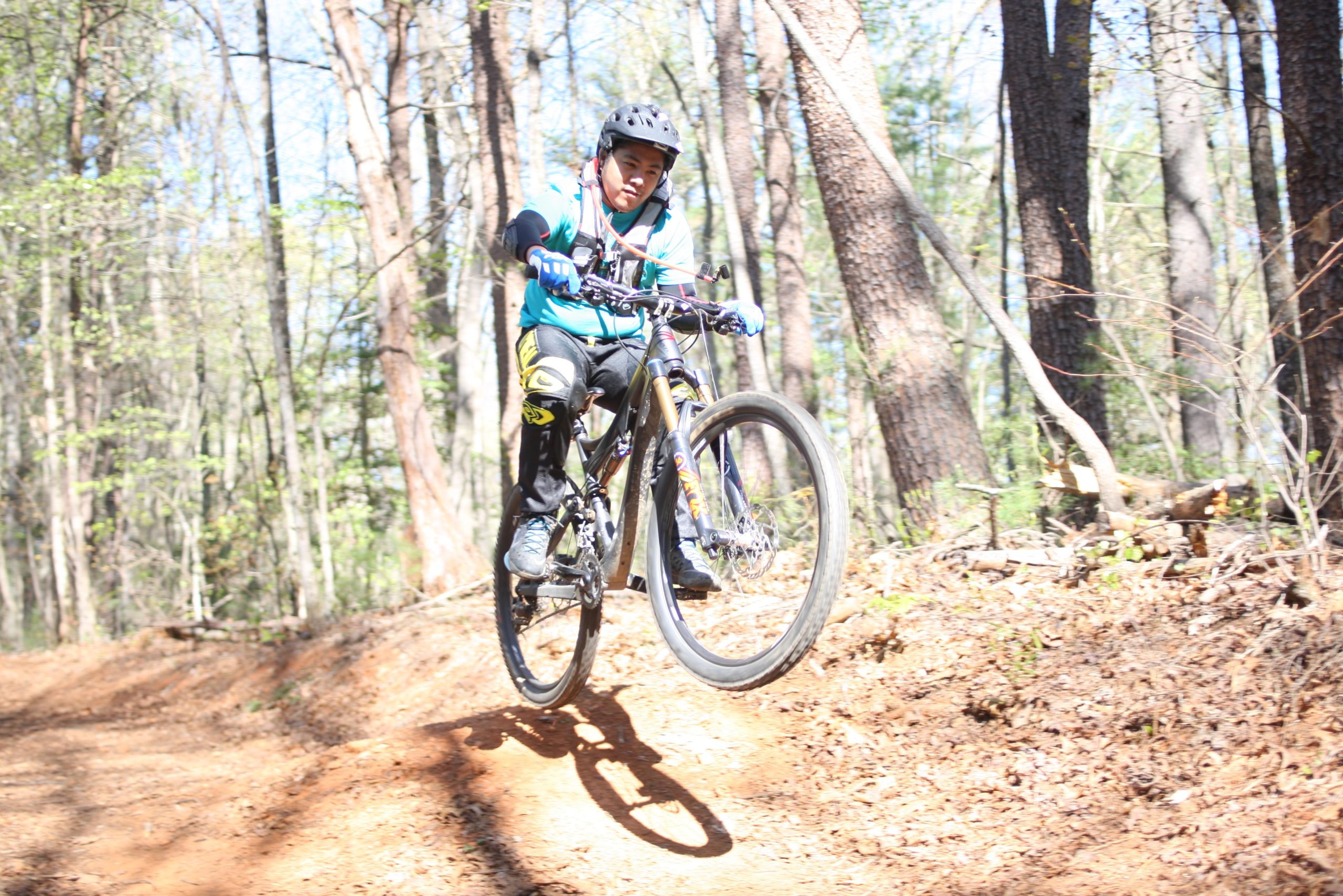 A young rider in a blue shirt and helmet performs a jump on a mountain bike on a forest trail, surrounded by trees and sunlight. Dark Mountain Trail mountain bike trail.