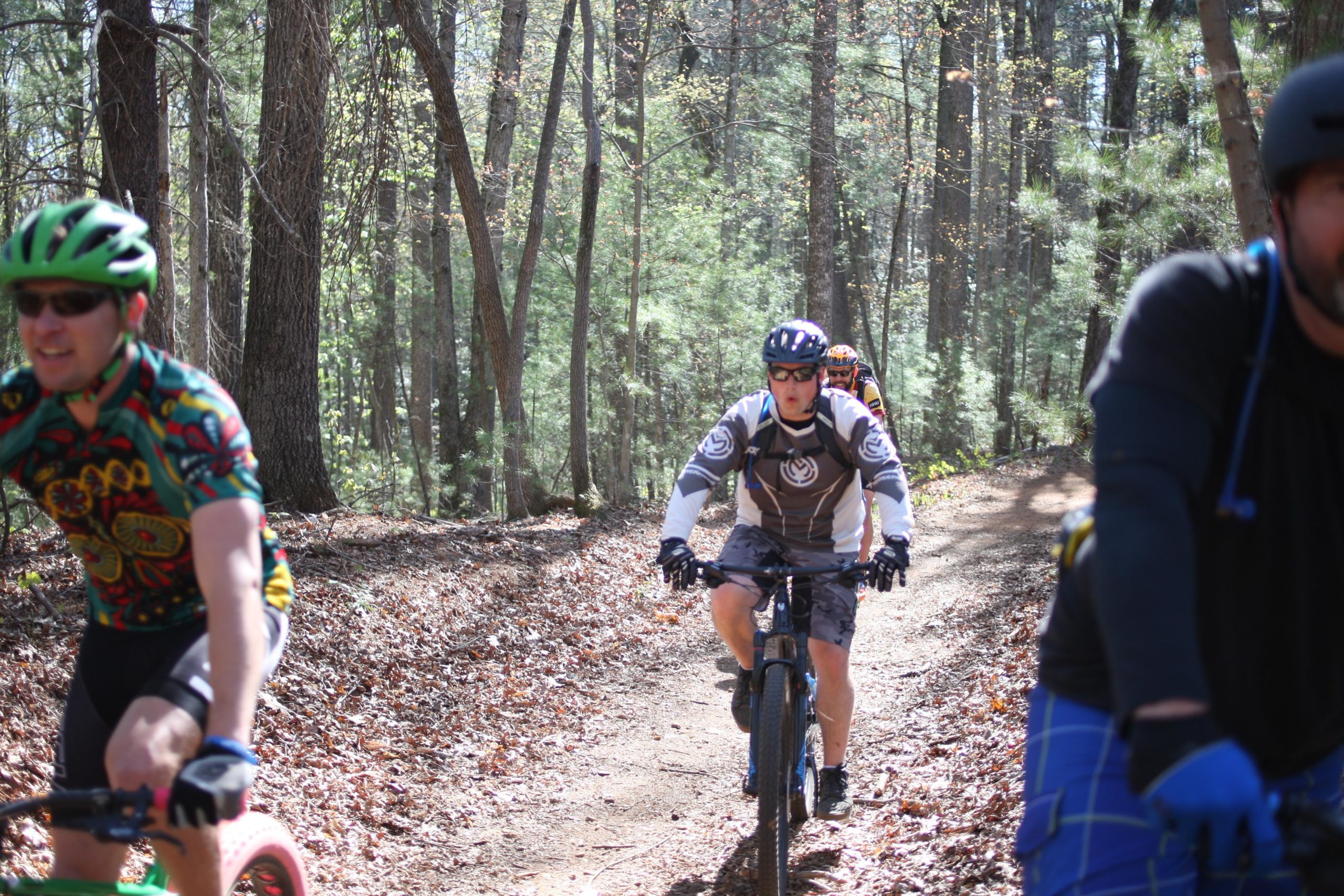 A group of mountain bikers riding on a sunlit forest trail, surrounded by trees and fallen leaves. The first rider is wearing a colorful patterned jersey and green helmet, while the second rider is in a gray and white outfit. The atmosphere conveys a sense of adventure and outdoor activity. Dark Mountain Trail mountain bike trail.
