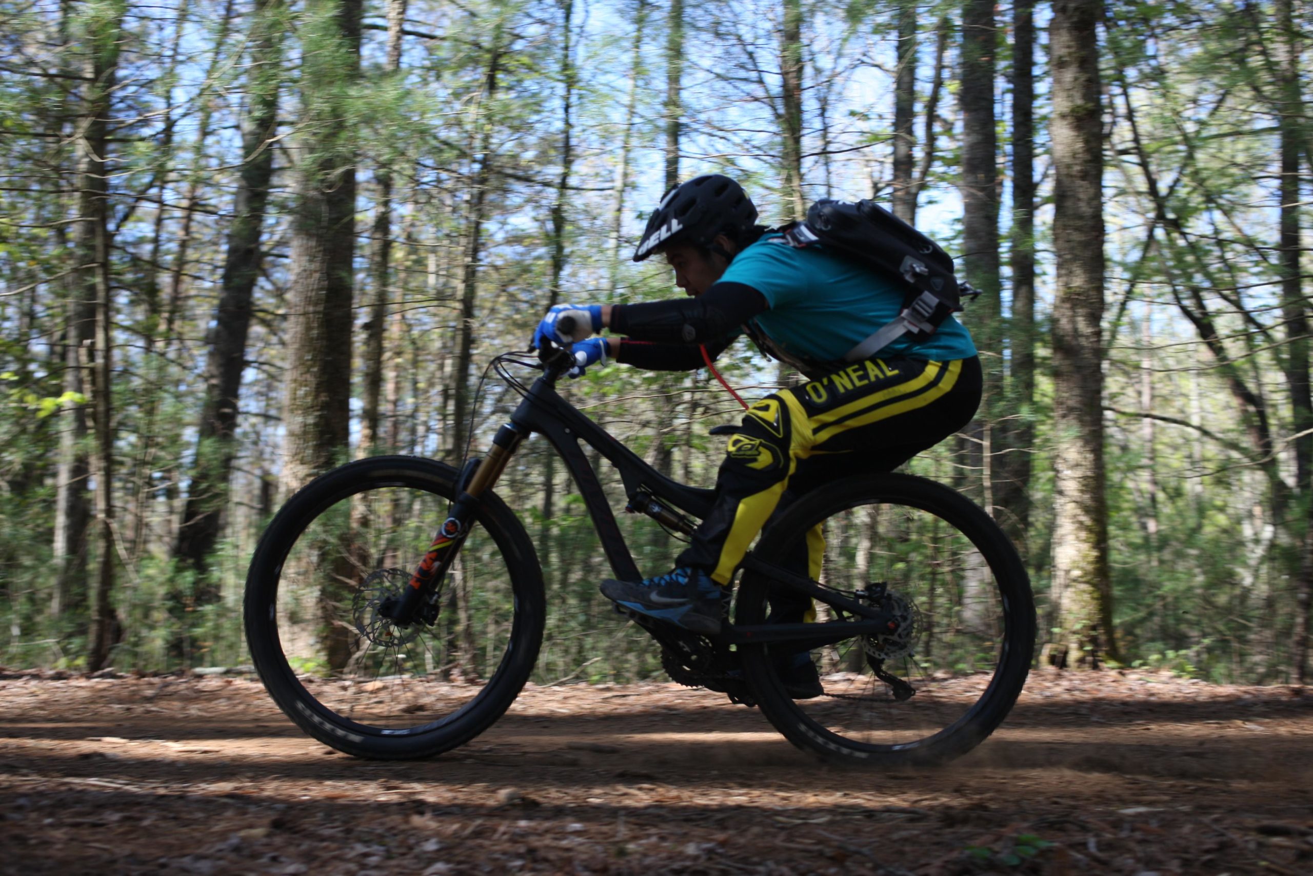 A mountain biker in a blue shirt and yellow pants rides swiftly along a dirt trail lined by trees, captured in motion to convey speed and agility. Dark Mountain Trail mountain bike trail.