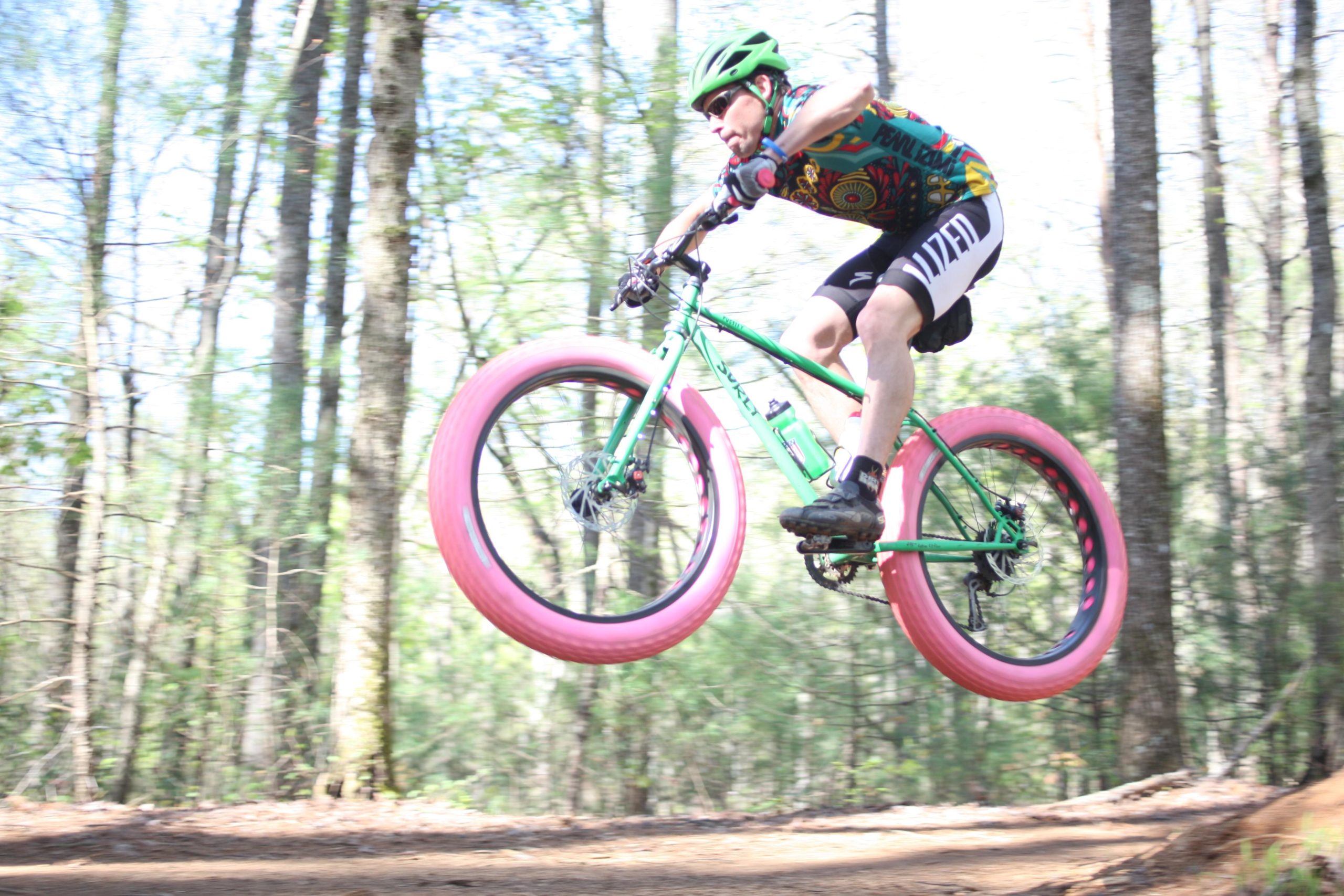 A cyclist performing a jump on a green bike with pink tires in a wooded trail, surrounded by trees and sunlight filtering through the leaves. Dark Mountain Trail mountain bike trail.