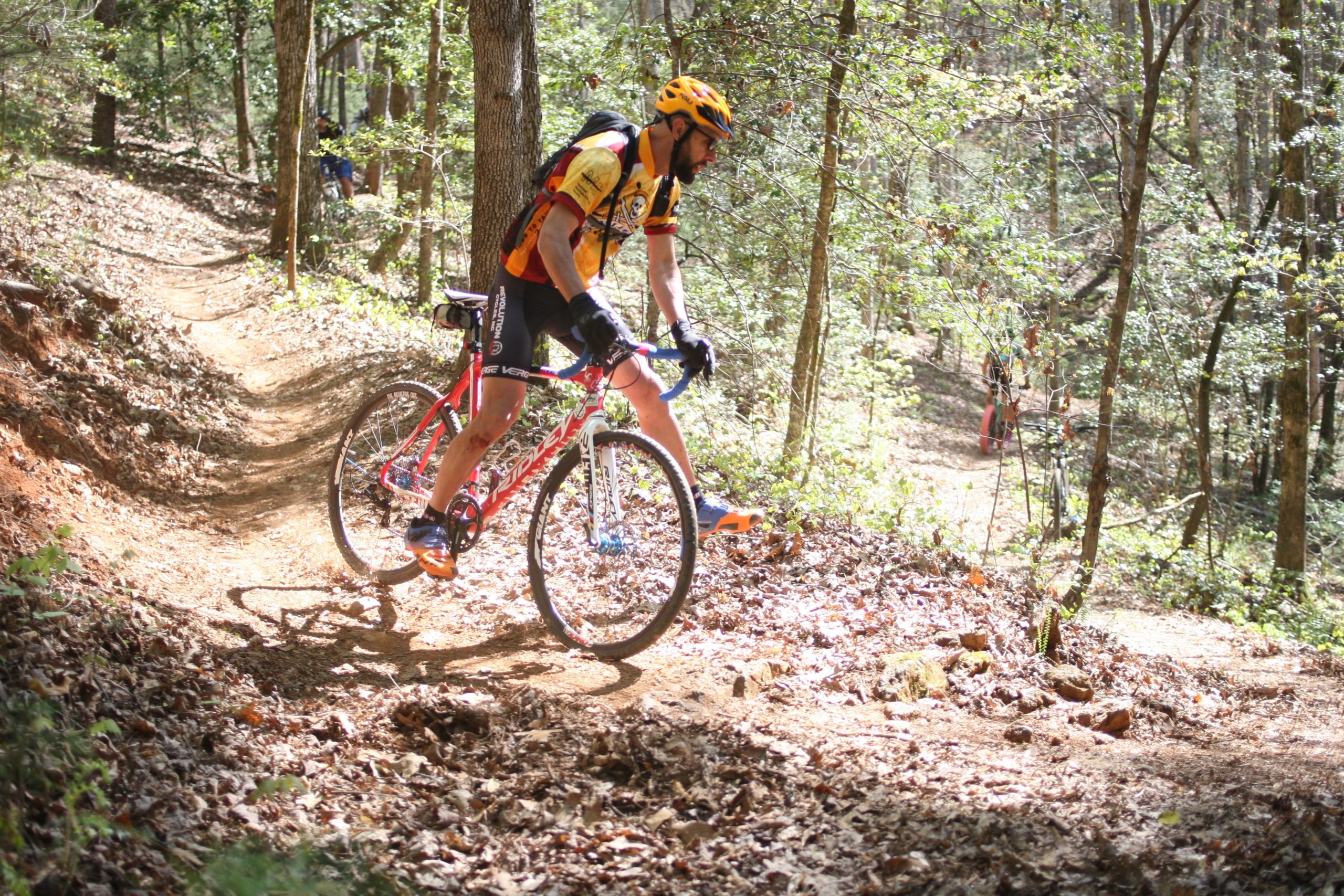 A cyclist riding a red bike on a dirt trail in a wooded area, surrounded by trees and foliage. Sunlight filters through the leaves, casting dappled shadows on the ground. The cyclist is wearing a yellow and orange jersey and a helmet, focusing intently on the path ahead. In the background, another cyclist can be seen riding on a similar trail. Dark Mountain Trail mountain bike trail.