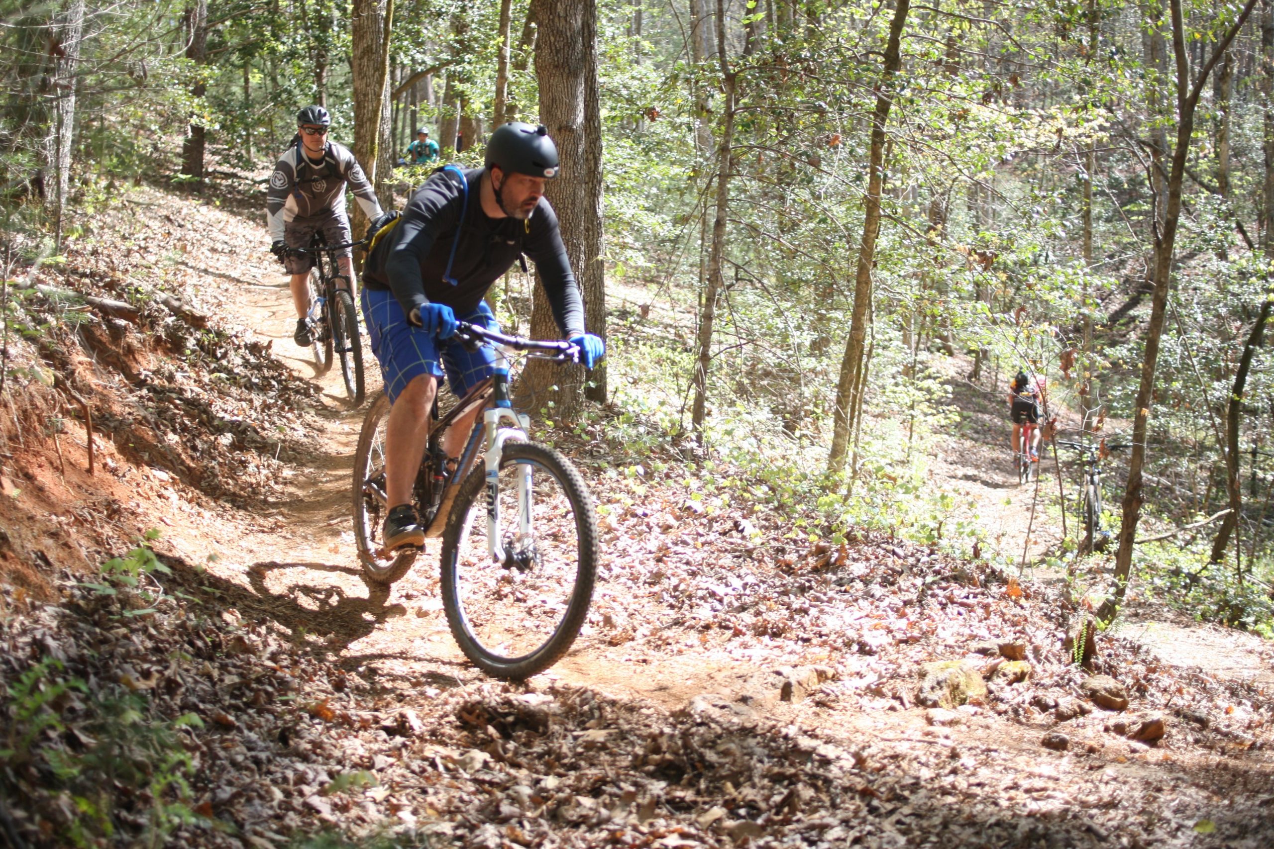 Three mountain bikers navigate a wooded trail, with one rider in the foreground wearing a black helmet and blue gloves, focusing on maneuvering their bike along the path. The trail is lined with trees and covered in fallen leaves, reflecting a sunny day outdoors. Dark Mountain Trail mountain bike trail.