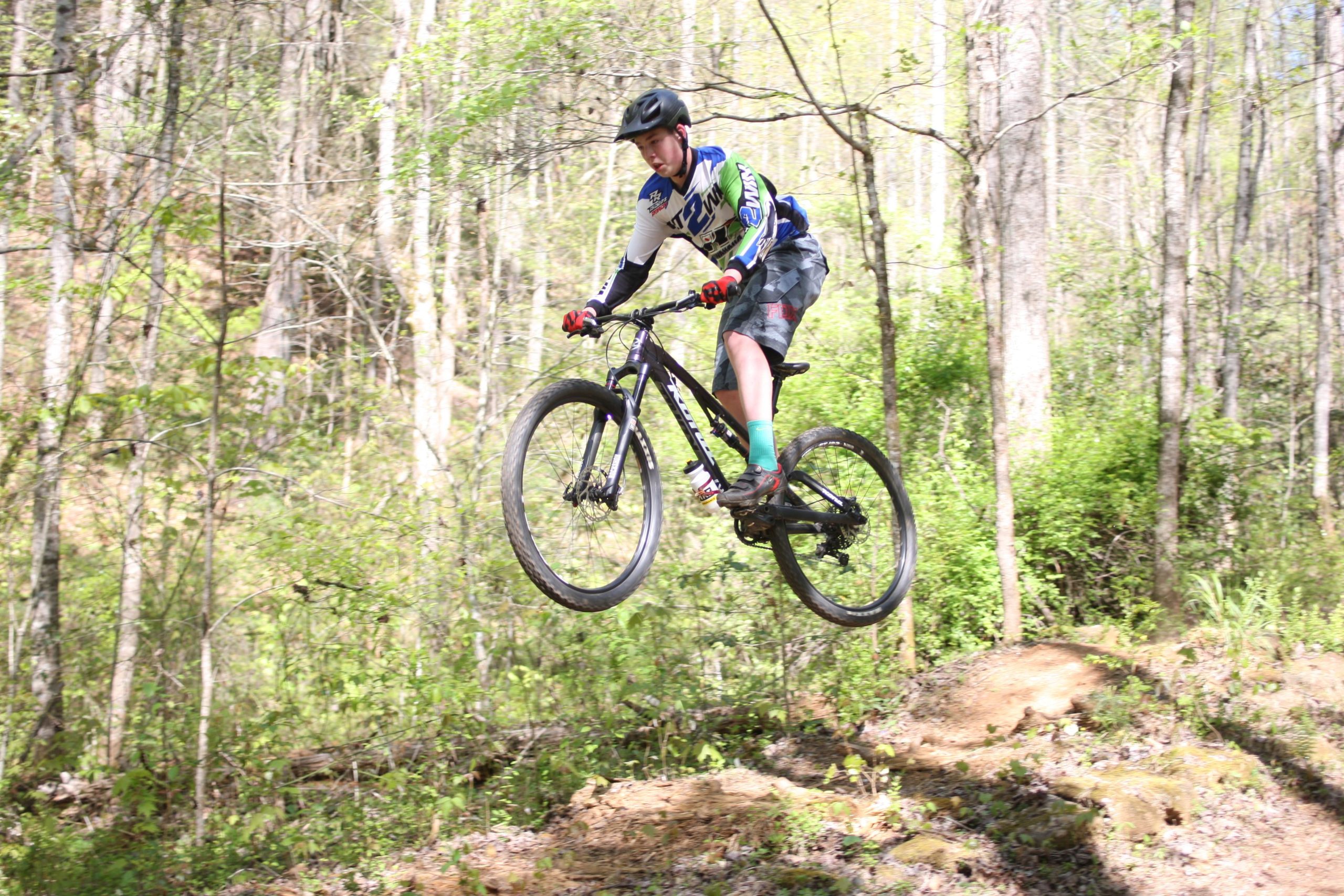 A young cyclist performing a jump on a mountain bike in a wooded trail, surrounded by greenery. The rider is wearing a helmet, gloves, and sports attire, showcasing a dynamic mid-air pose as the bike's front wheel is elevated off the ground. Dark Mountain Trail mountain bike trail.