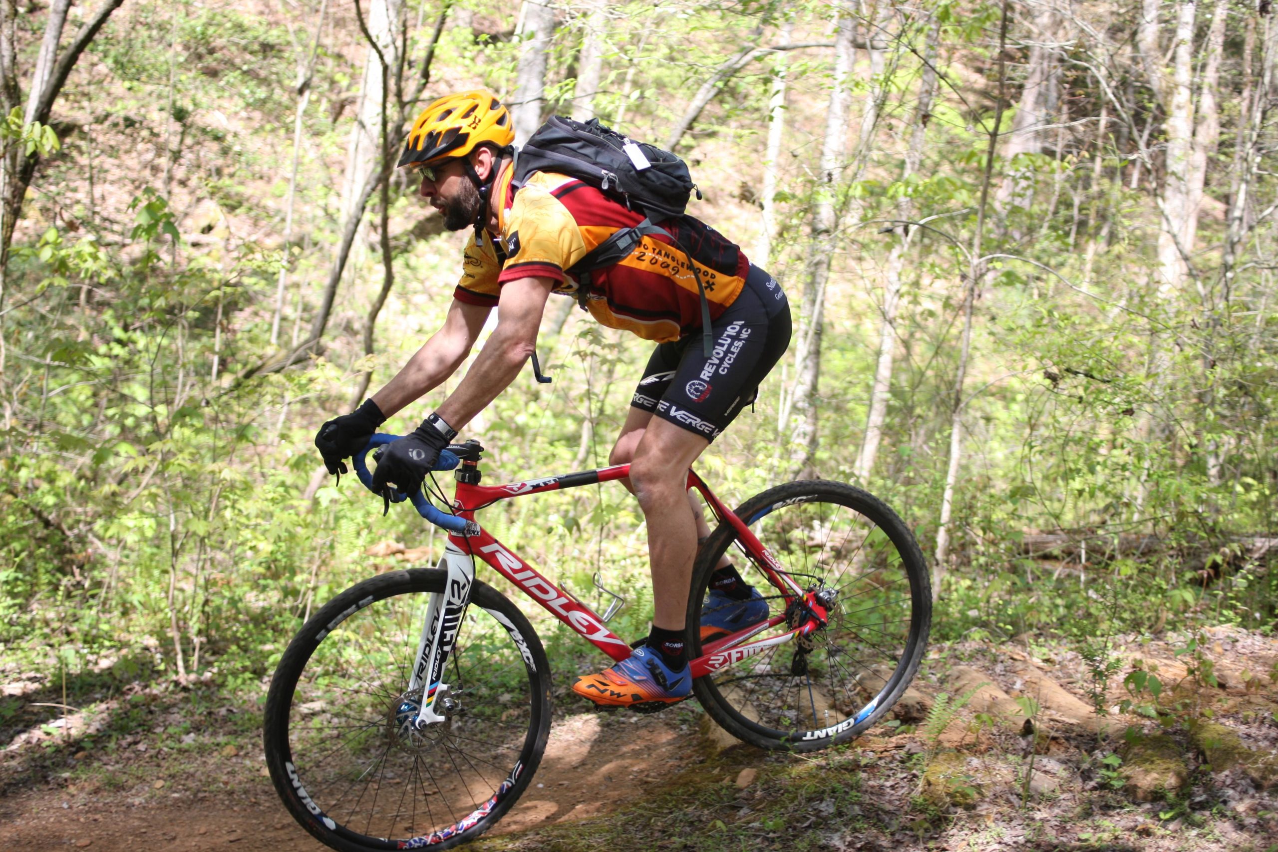 A cyclist riding a red mountain bike on a trail surrounded by greenery, wearing a helmet and sporty attire. The cyclist is focused on navigating the path, with trees and lush foliage in the background. Dark Mountain Trail mountain bike trail.