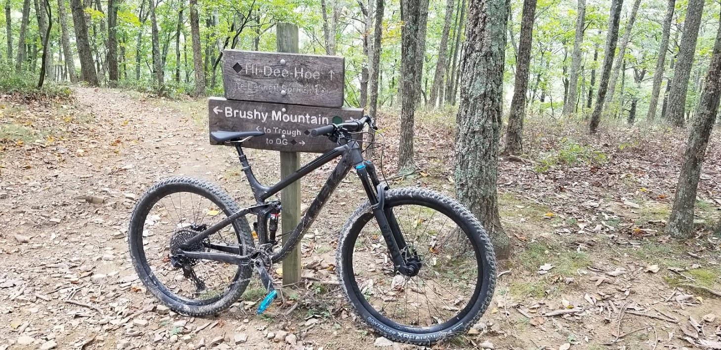 A black mountain bike leaning against a wooden trail sign in a wooded area. The sign points in two directions: one towards "Hi Dee Ho" and the other towards "Brushy Mountain." The ground is covered with leaves and dirt, surrounded by trees with green foliage. Carvin's Cove Trail system mountain bike trail.