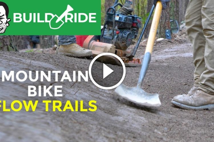 A close-up shot of a person using a shovel to shape a dirt surface while working on mountain bike flow trails. In the background, a small gas-powered tiller is visible, along with trees and natural surroundings. The image includes the text "Build Ride Mountain Bike Flow Trails" at the top.