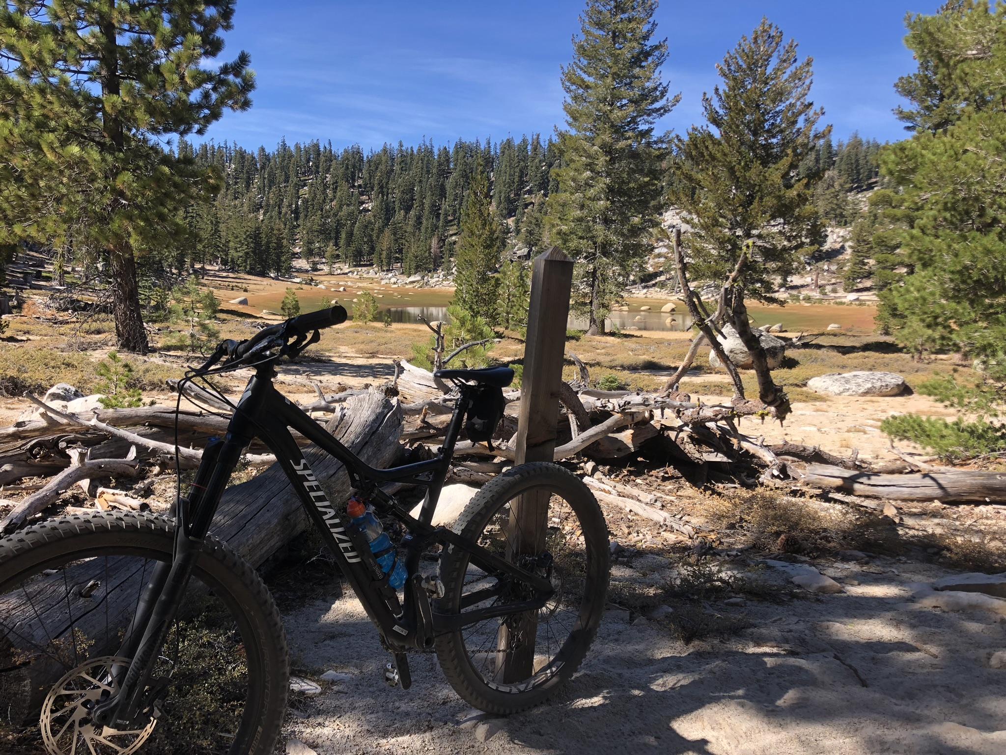 A mountain bike is parked on a rocky trail, surrounded by a scenic landscape of pine trees and a small lake in the background. The area is dotted with logs and boulders, under a clear blue sky. Tahoe Rim Trail: Tahoe Meadows to Tunnel Creek Road / Flume Trail mountain bike trail.