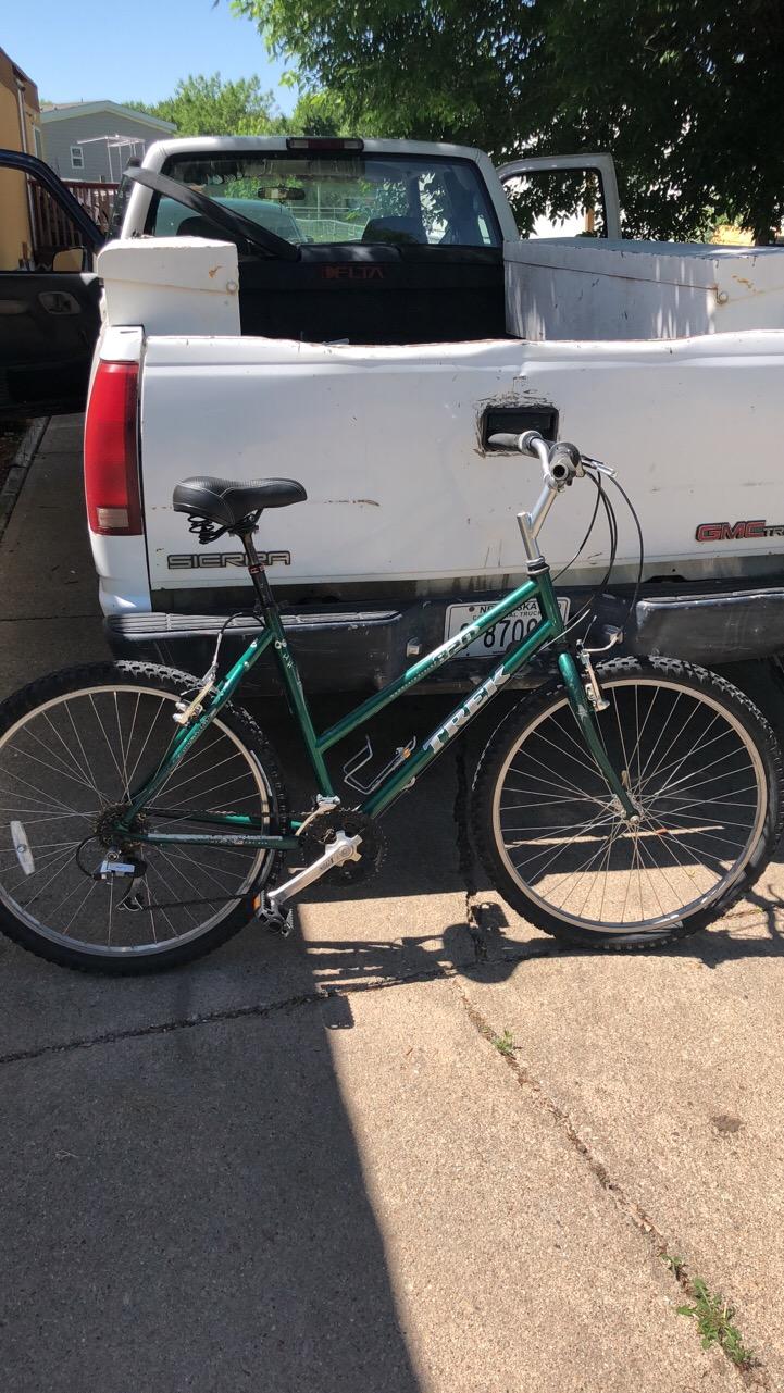 Trek 820: A green Trek mountain bike resting against the back of a white pickup truck, with a sunlit background and shadows cast on the pavement. The truck's bed is open, revealing some tools or equipment inside.