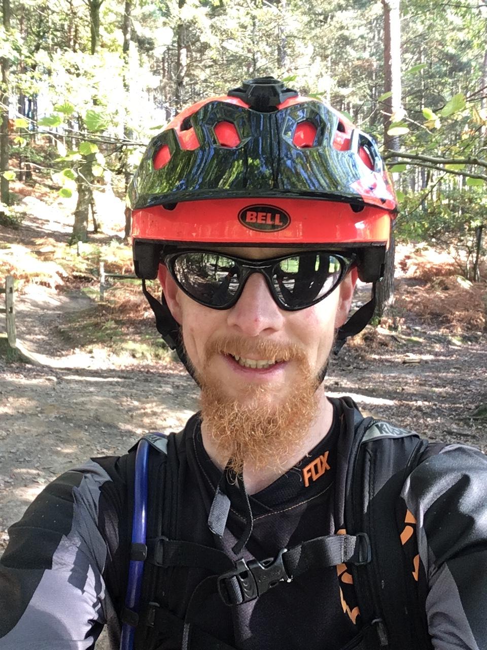 A man wearing a bright orange and black mountain biking helmet and sunglasses is smiling at the camera. He is dressed in a black long-sleeve shirt with orange accents and is standing in a wooded area with a dirt path visible behind him. The scene is well-lit, showcasing lush green trees and foliage in the background. Barry Knows Best mountain bike trail.