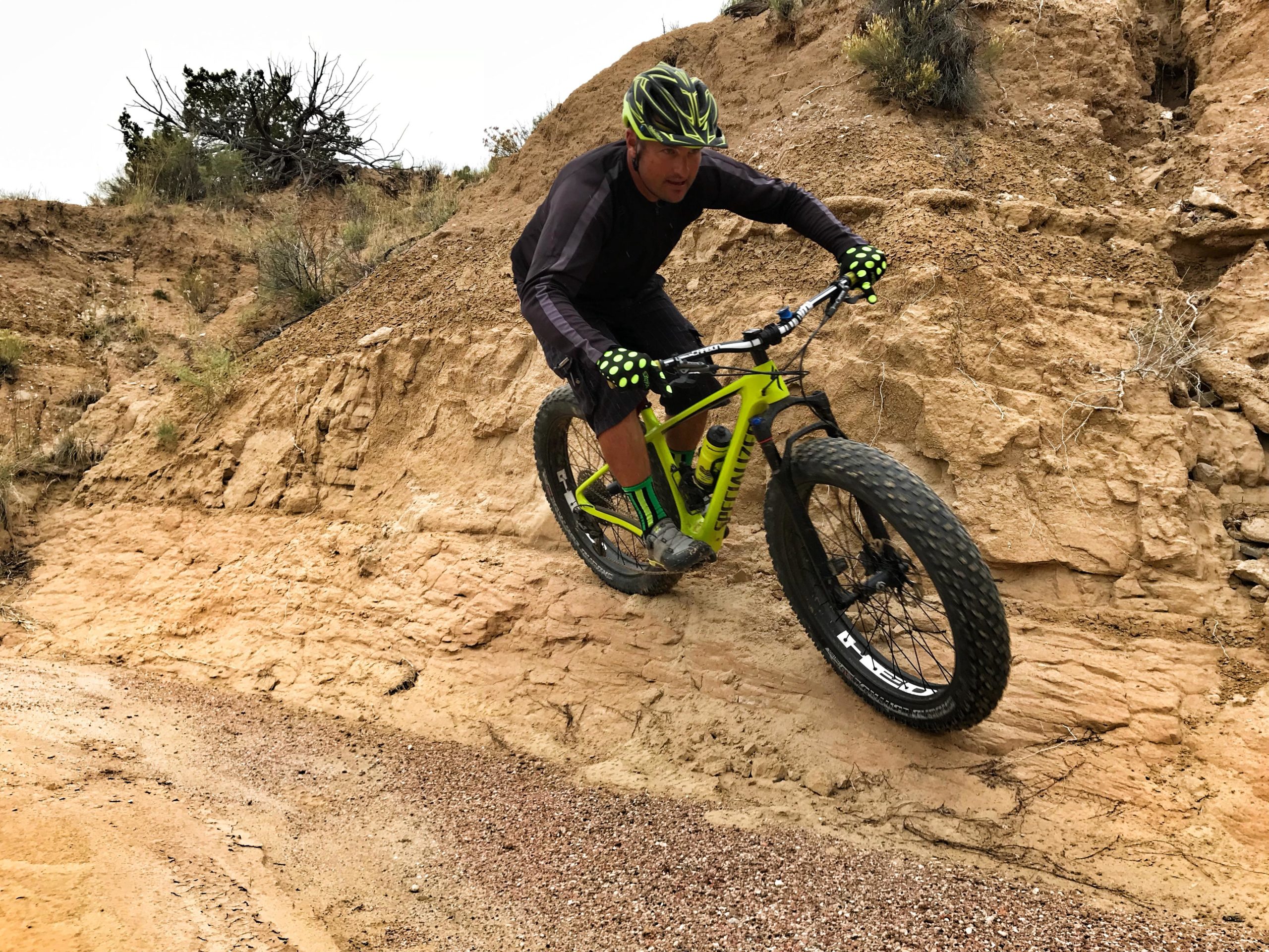 A man in athletic wear rides a bright green fat bike on a dirt trail, navigating a steep, sandy incline. He wears a helmet and gloves for safety, showcasing an action-packed moment as he balances skillfully on the bike. The surrounding terrain is rocky and earthy, with sparse vegetation in the background. Mariposa Fat Bike Trails mountain bike trail.