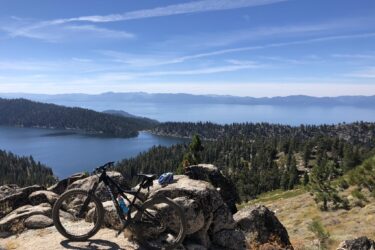 Mountain bike resting on rocks with a stunning view of a lake and forested mountains under a clear blue sky. Tahoe Rim Trail: Tahoe Meadows to Tunnel Creek Road / Flume Trail mountain bike trail.
