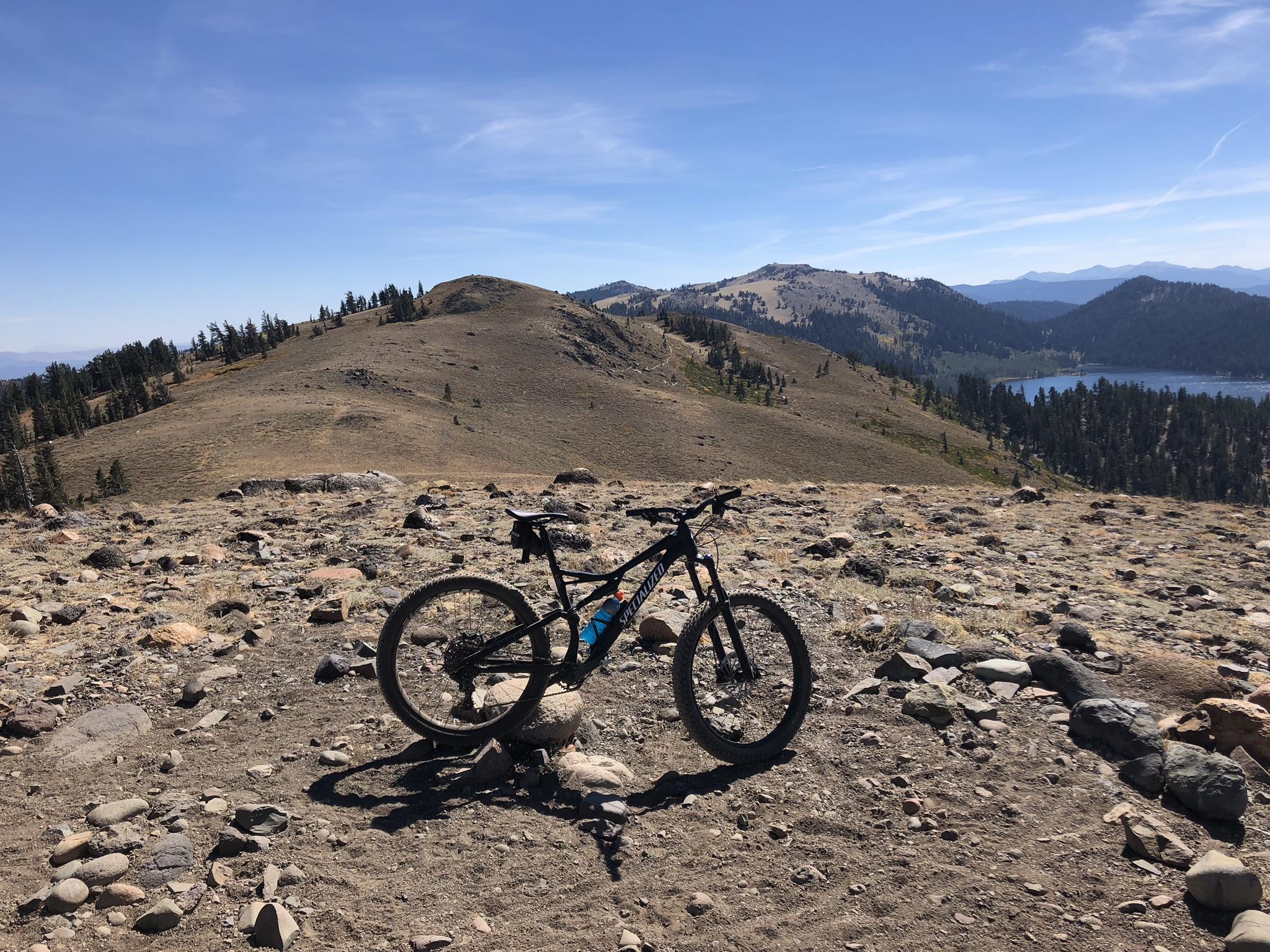 A mountain bike rests on rocky terrain with a panoramic view of rolling hills and a lake in the background, under a clear blue sky. Tahoe Rim Trail: Tahoe Meadows to Tunnel Creek Road / Flume Trail mountain bike trail.
