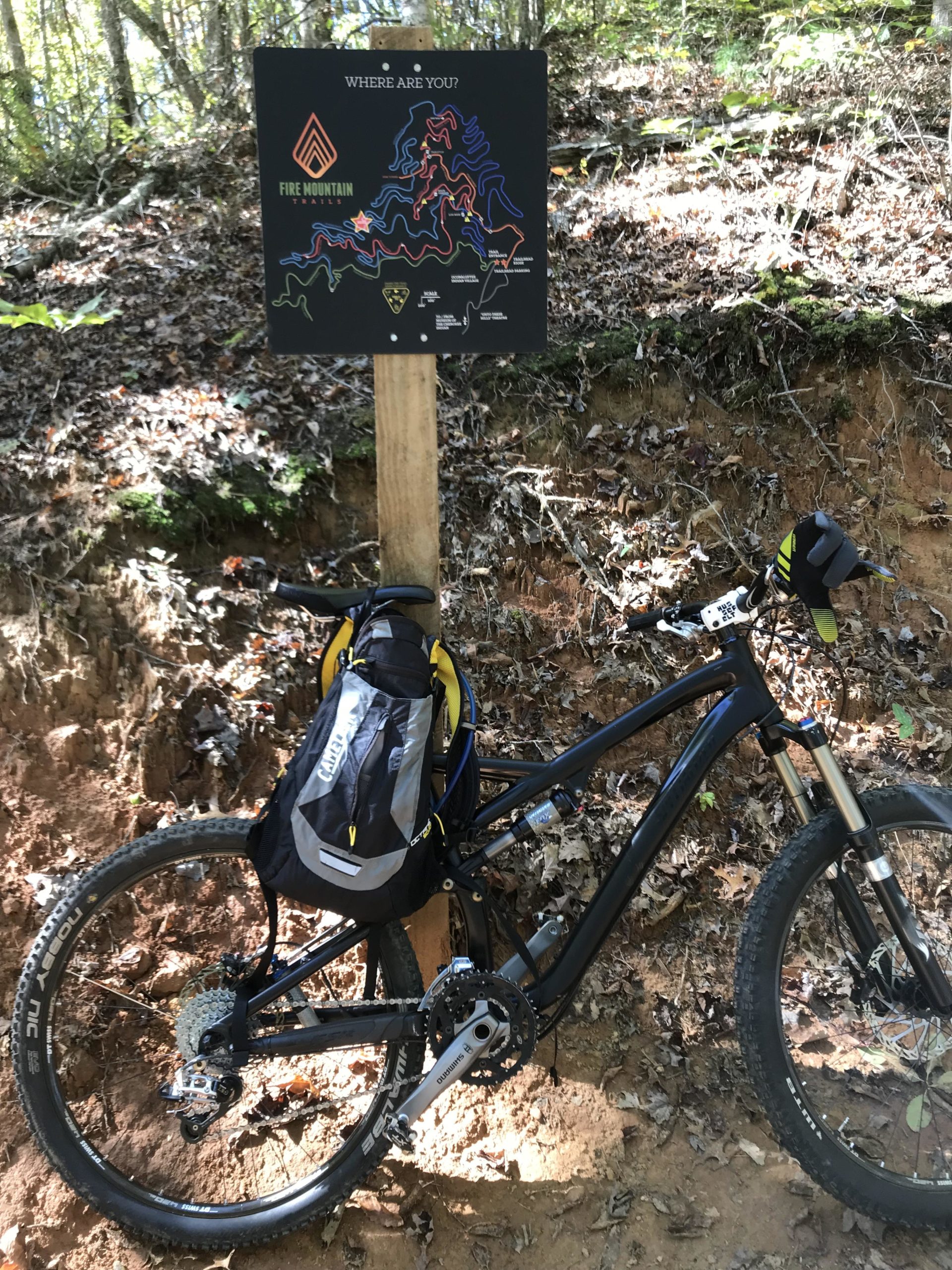 A mountain bike with a backpack rests on the ground next to a trail map sign for Fire Mountain Trails, surrounded by autumn foliage. The sign features a colorful map showing various biking trails in the area. Fire Mountain Trail System mountain bike trail.