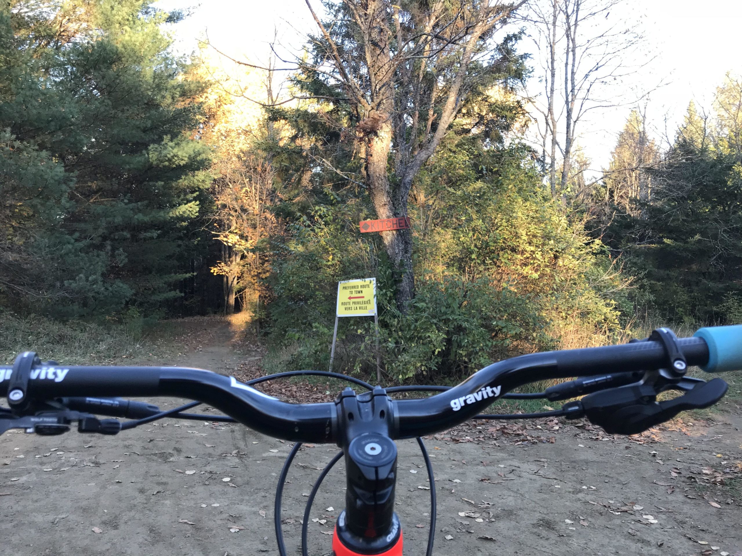 A view from the handlebars of a mountain bike, looking down a dirt path surrounded by trees. In the foreground, the bike's handlebars and grips are visible, with a sign indicating a trail closure on the right side. The path ahead leads into a wooded area with autumn foliage. Kingdom Trails mountain bike trail.