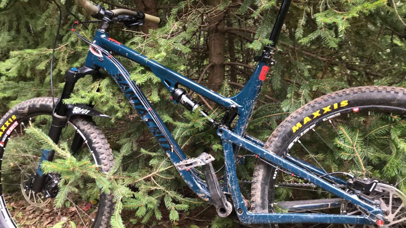 Santa Cruz 5010: A blue mountain bike partially obscured by greenery, showing its rugged tires and front suspension. The bike appears to have dirt and mud on it, suggesting recent use on a trail or outdoor adventure.