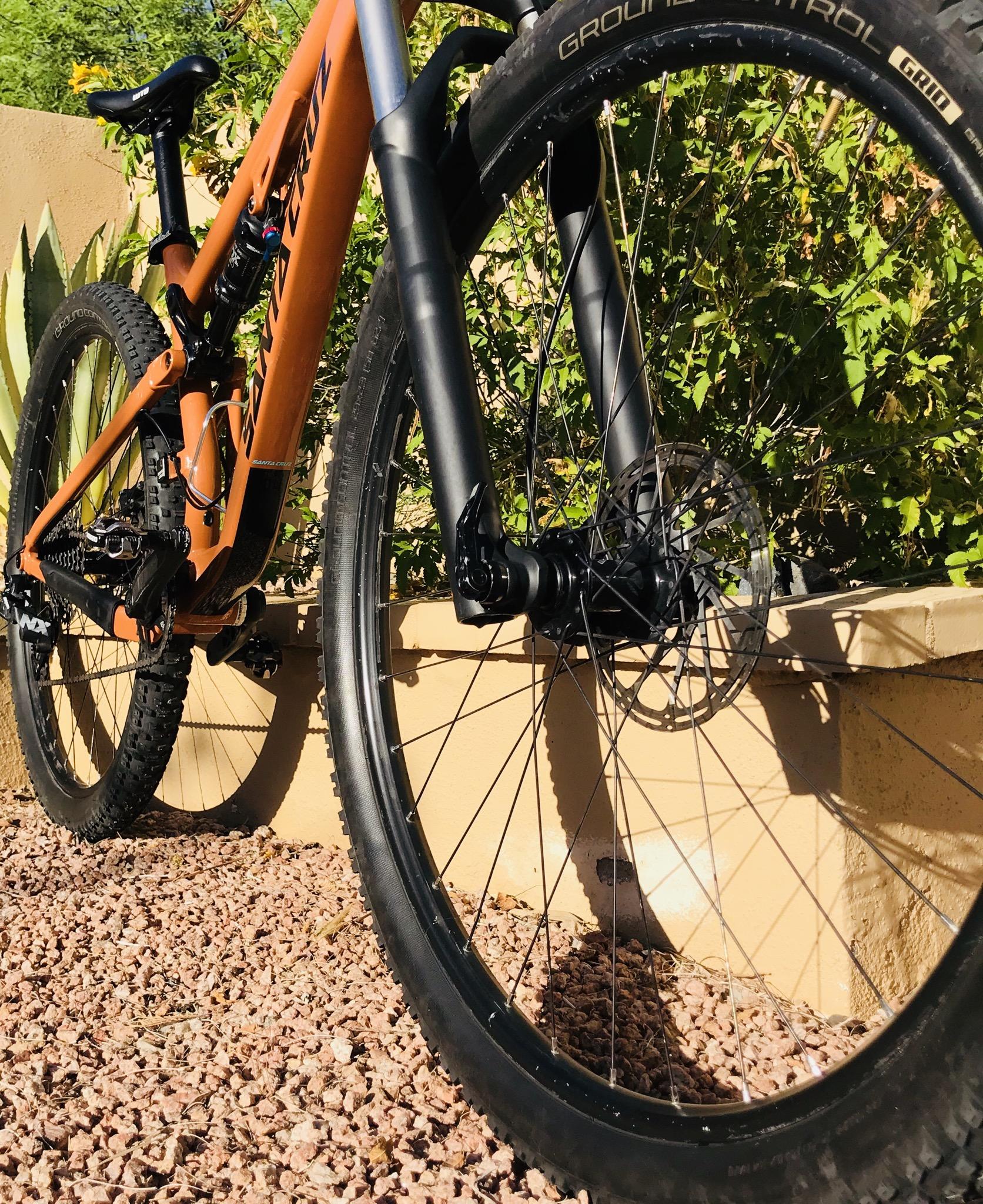 Santa Cruz Tallboy: A close-up view of the front wheel and frame of an orange mountain bike, positioned against a backdrop of green foliage and a light-colored wall. The wheel features thick, knobby tires suitable for off-road terrain, and a detailed look at the disc brake and fork suspension components is visible. The scene is set on rocky ground with pebbles.