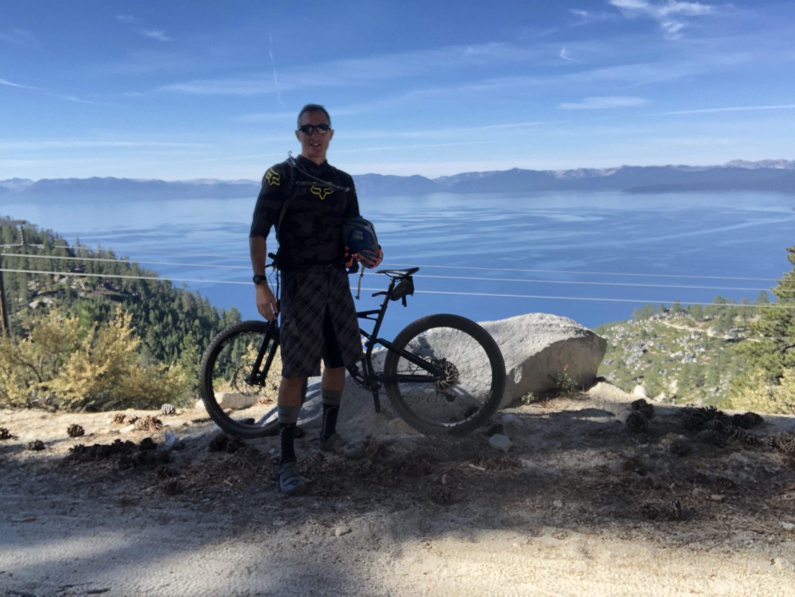 A mountain biker stands next to a bicycle on a rocky trail overlooking a scenic lake surrounded by trees and mountains. The sky is clear with a few clouds, and the water reflects the blue hues of the sky. The biker is wearing sunglasses, a black and yellow jersey, and plaid shorts. Flume Trail mountain bike trail.