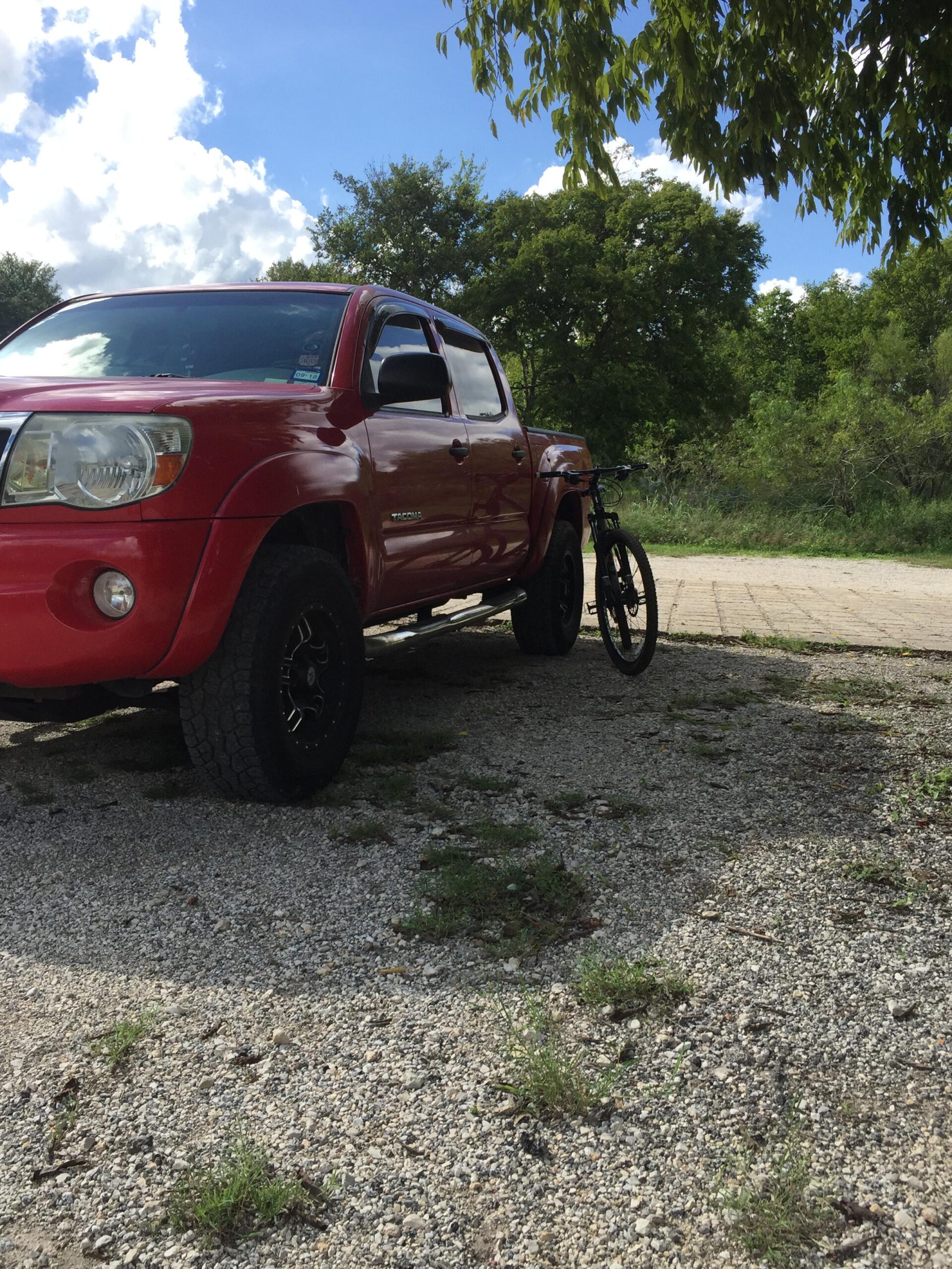 Trek Trek Marlin 4 (29er): Red pickup truck parked on a gravel surface, with a bicycle leaning against it. Lush greenery and a blue sky with fluffy clouds in the background.