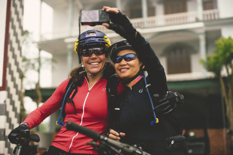 Two women in cycling gear pose for a selfie, smiling broadly while wearing helmets and sunglasses. They are standing next to their bicycles, with an outdoor setting featuring a building in the background.