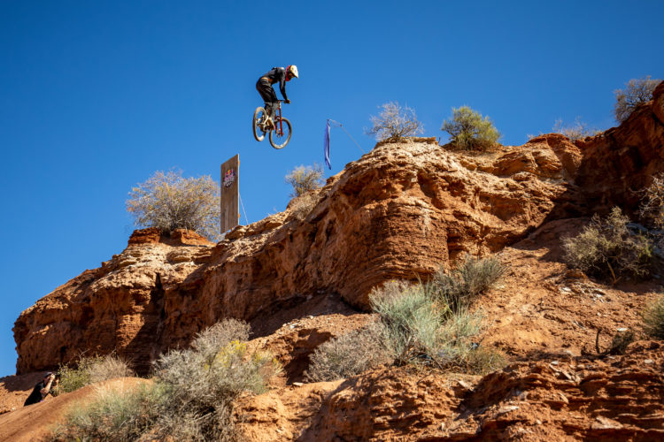 A mountain biker performing a jump off a rocky cliff against a clear blue sky, with desert vegetation and a wooden ramp in the background.