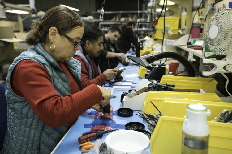A group of workers engaged in assembly tasks at a workspace filled with tools and components. In the foreground, a woman focuses on her work while holding a tool, wearing glasses and a blue vest. Other workers are seated at the table, each involved in similar activities, surrounded by yellow storage bins and various items needed for their tasks. The setting appears to be a factory or workshop environment.