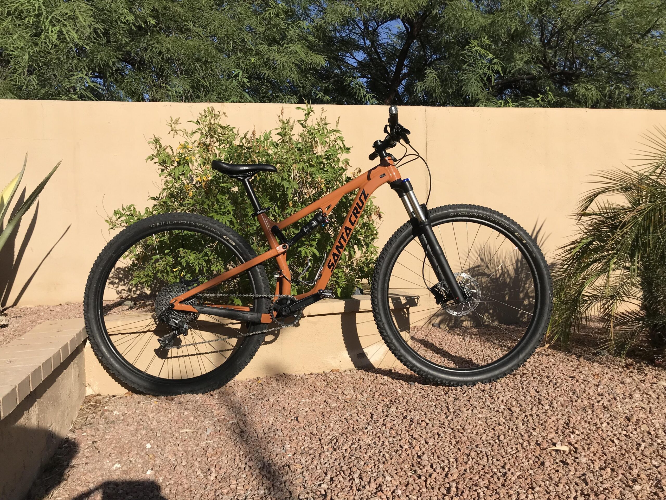Santa Cruz Tallboy: A mountain bike with an orange frame is leaning against a light-colored wall, surrounded by greenery and gravel. The bike features thick tires, a front suspension fork, and a single seat. Sunlight casts shadows on the ground, highlighting the bike's design and the natural surroundings.