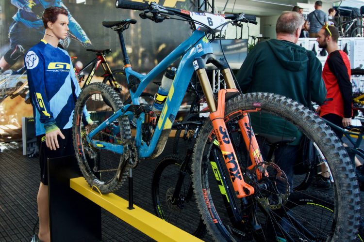 A blue mountain bike displayed on a yellow stand, featuring a mannequin in a blue cycling jersey alongside it. In the background, attendees are engaged in conversations at a biking event, with promotional graphics visible.