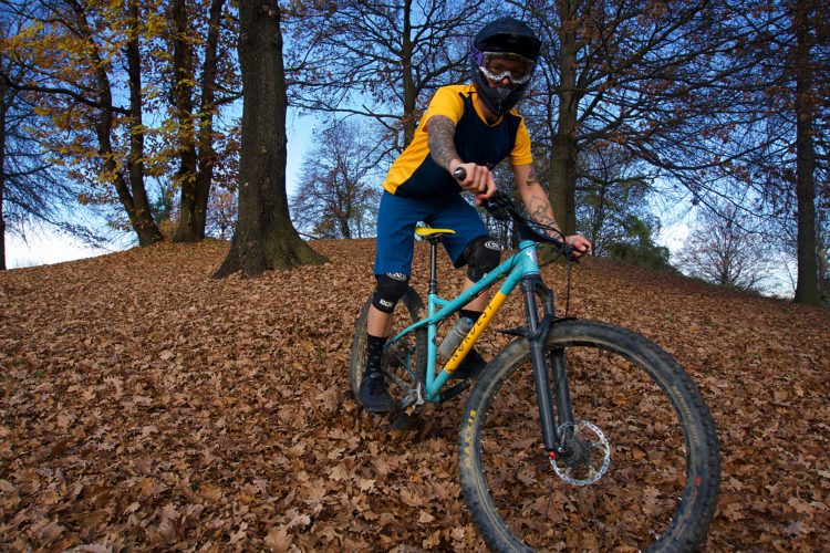 A mountain biker wearing a helmet and protective gear rides through a landscape covered in autumn leaves, with trees and a blue sky in the background. The biker is positioned on a sloped area, showcasing the fall season's vibrant foliage.