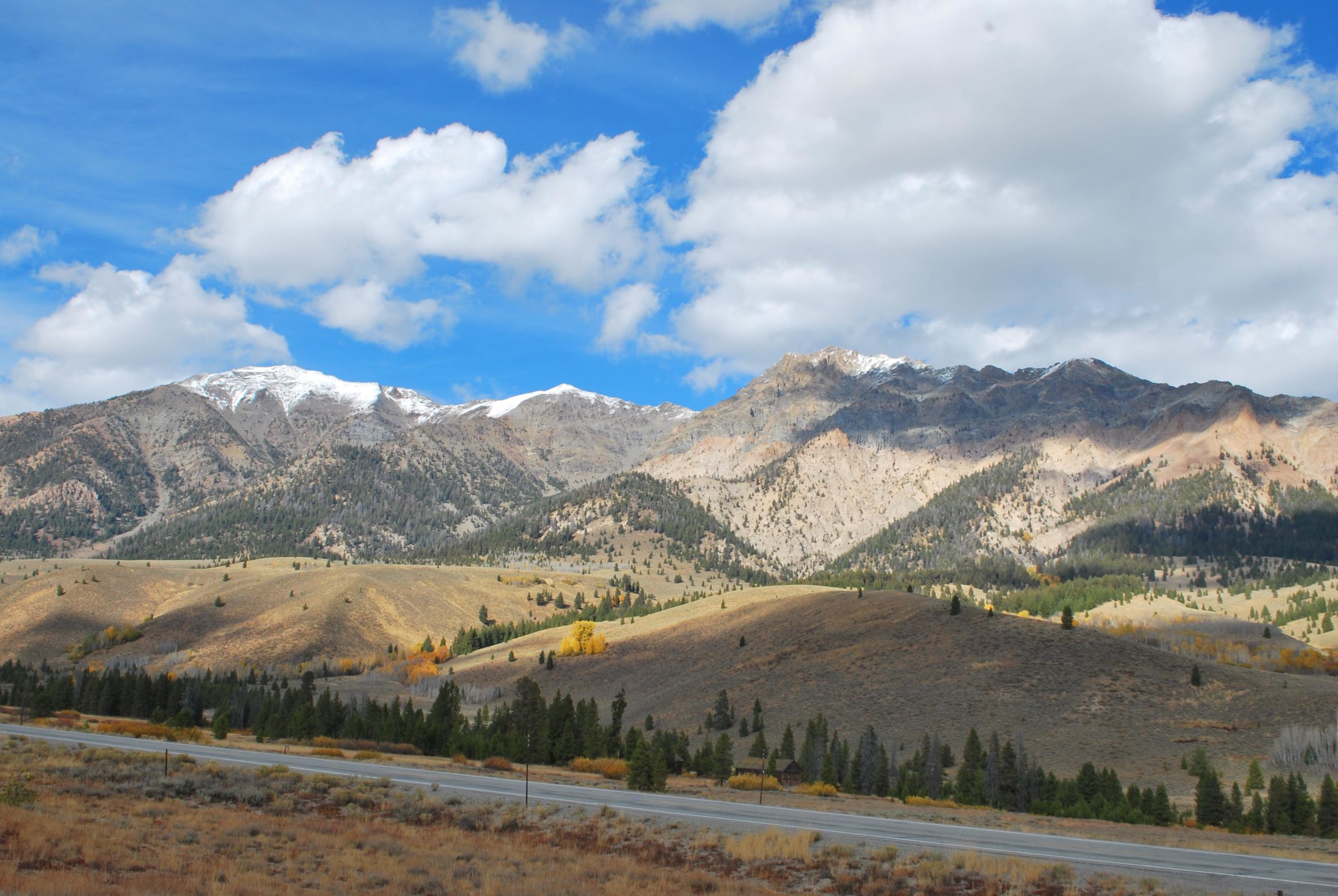 A scenic view of mountains with snow-capped peaks under a partly cloudy blue sky. In the foreground, rolling hills covered in shrubs and patches of green trees are visible, along with a winding road. The landscape showcases a mix of colors, highlighting the natural beauty of the area. Harriman Trail mountain bike trail.