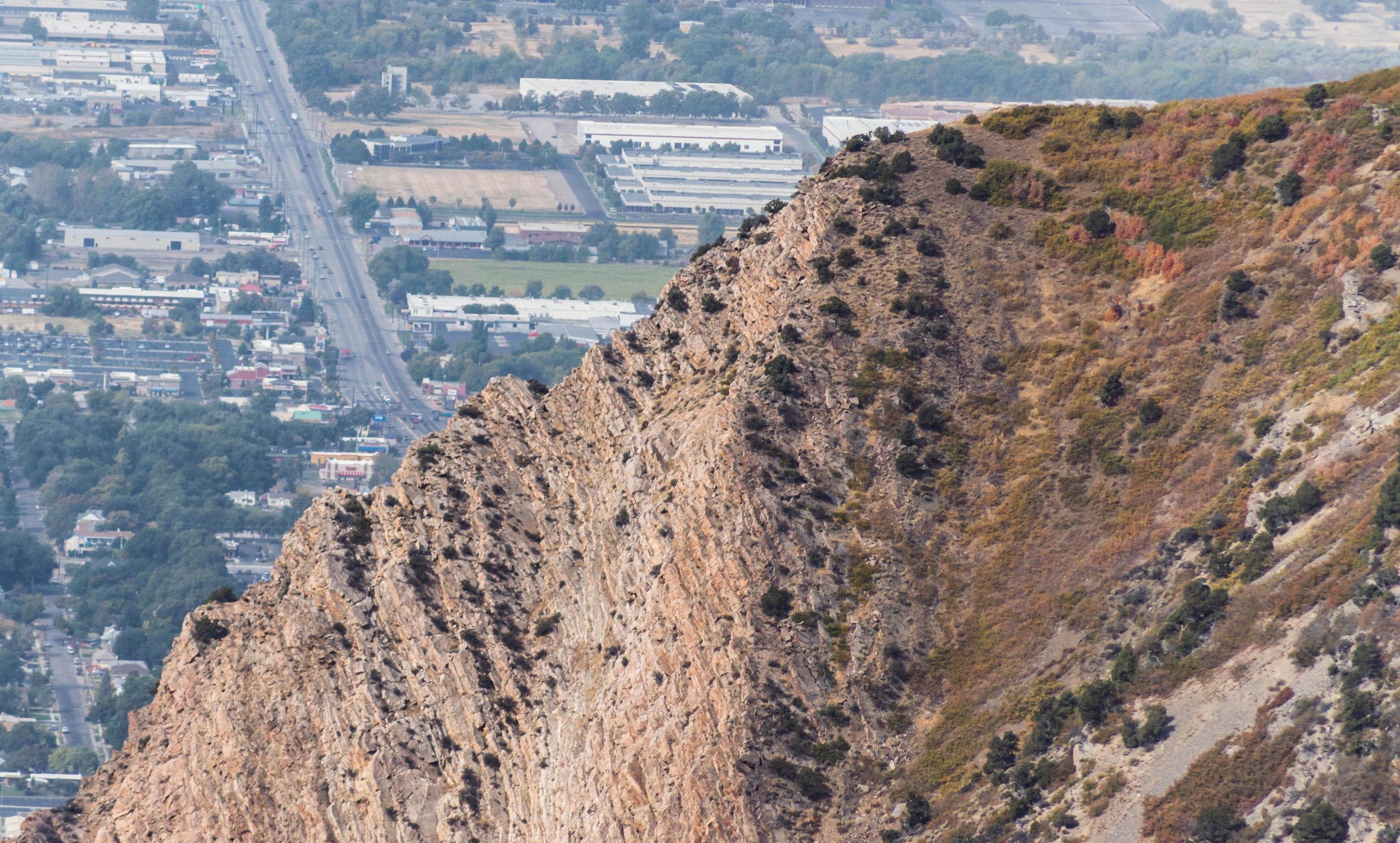 A rocky mountain ridge with steep slopes in the foreground, showing textured geological formations. In the background, a view of a cityscape with roads, buildings, and greenery along the outskirts, indicating a blend of natural and urban settings. Sardine Peak Loop mountain bike trail.