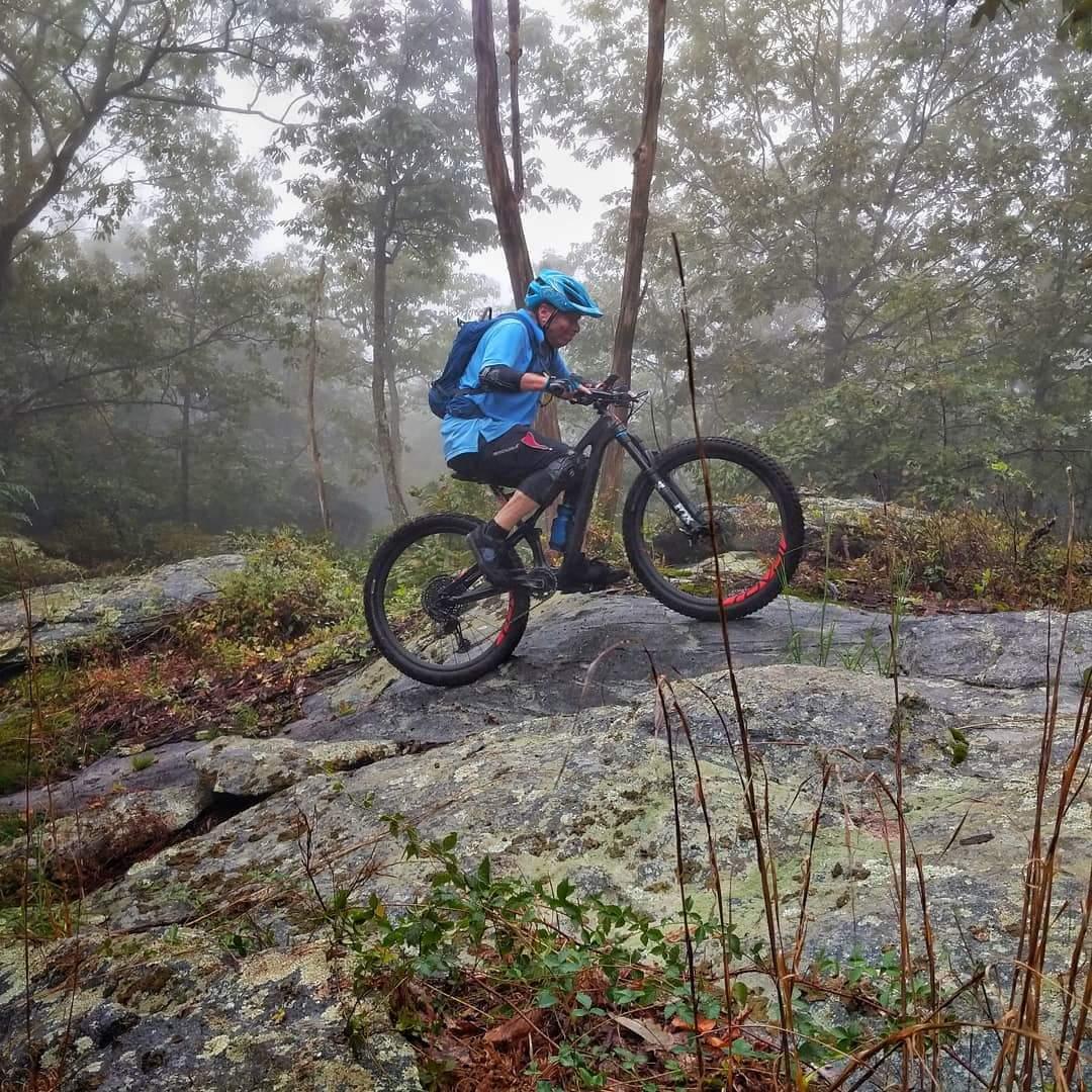 A mountain biker navigating rocky terrain in a misty forest. The rider, wearing a blue helmet and blue jersey, is mid-action, climbing over a large boulder surrounded by lush green vegetation and tall trees. The scene captures the adventurous spirit of mountain biking in a natural setting. Frederick Watershed mountain bike trail.