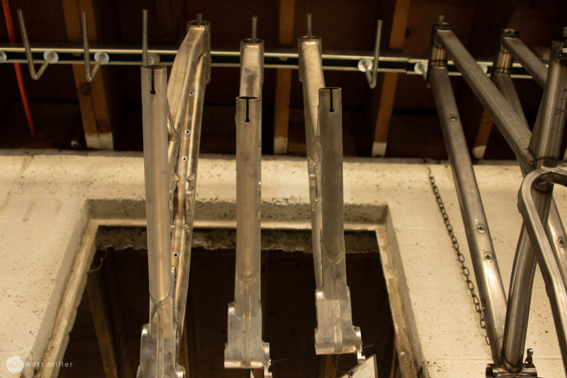 Aluminum bike frames hanging in a workshop, showcasing detailed craftsmanship and unfinished surfaces. The beams are positioned vertically against a textured wall with visible hooks and wooden beams overhead. A partially visible doorway is located at the bottom of the image.