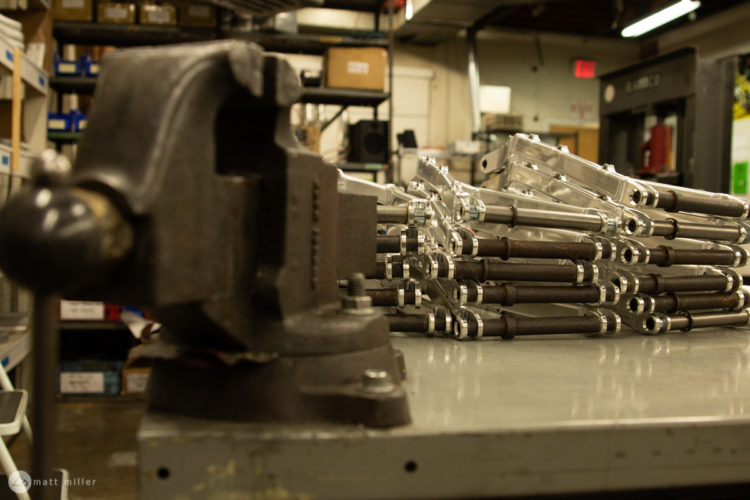 A close-up view of a black bench vise positioned in the foreground, with a stack of metallic components featuring silver and brown elements arranged neatly in the background. The setting appears to be a workshop or industrial space, with shelves filled with various materials visible.