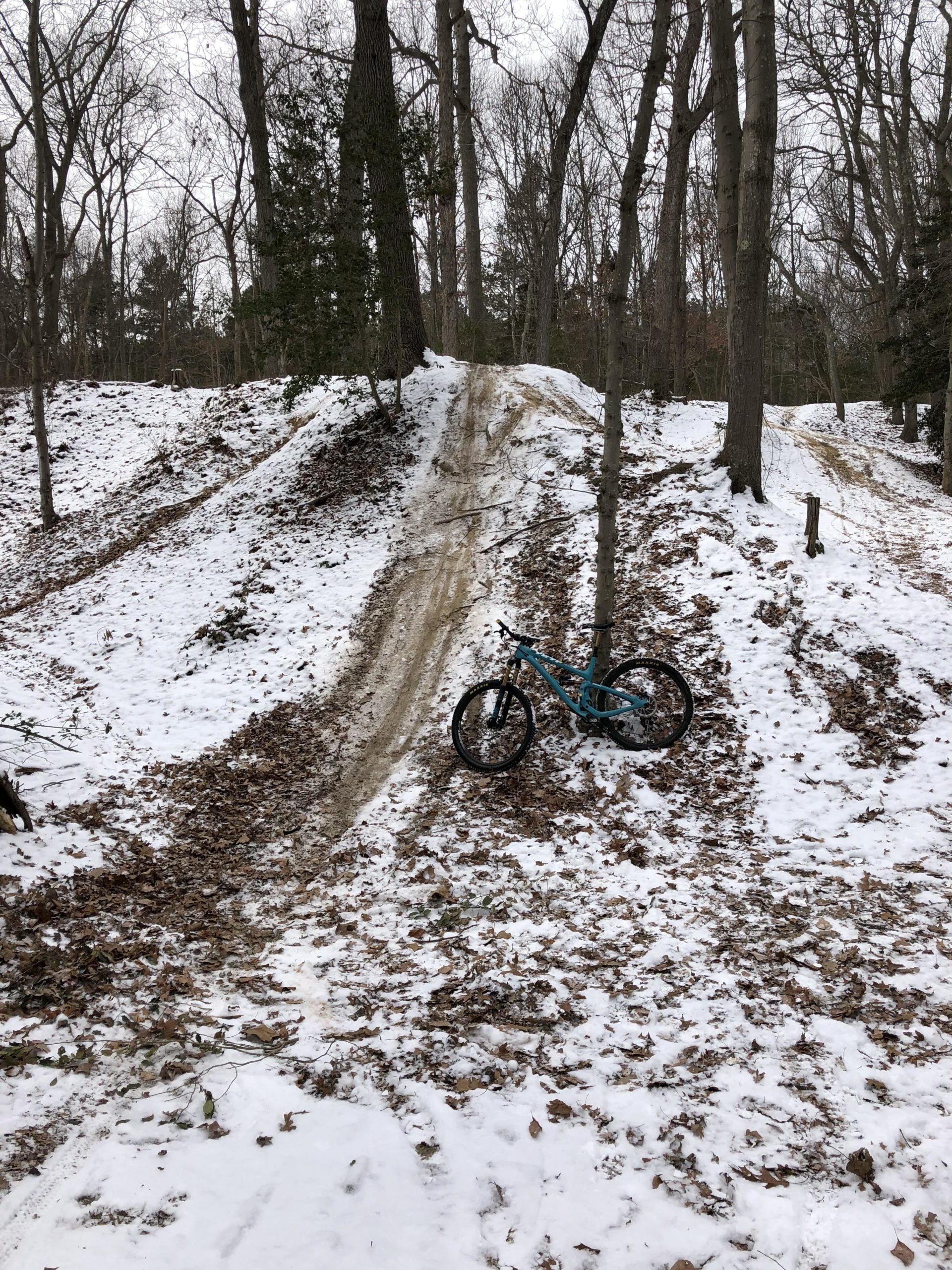 A mountain bike rests on the snowy, leaf-covered ground beside a dirt trail that leads up a hill in a wooded area. The scene features bare trees and patches of snow, indicating winter conditions. The bike is leaning against a tree, with a dirt track visible leading up the slope. Allaire State Park mountain bike trail.