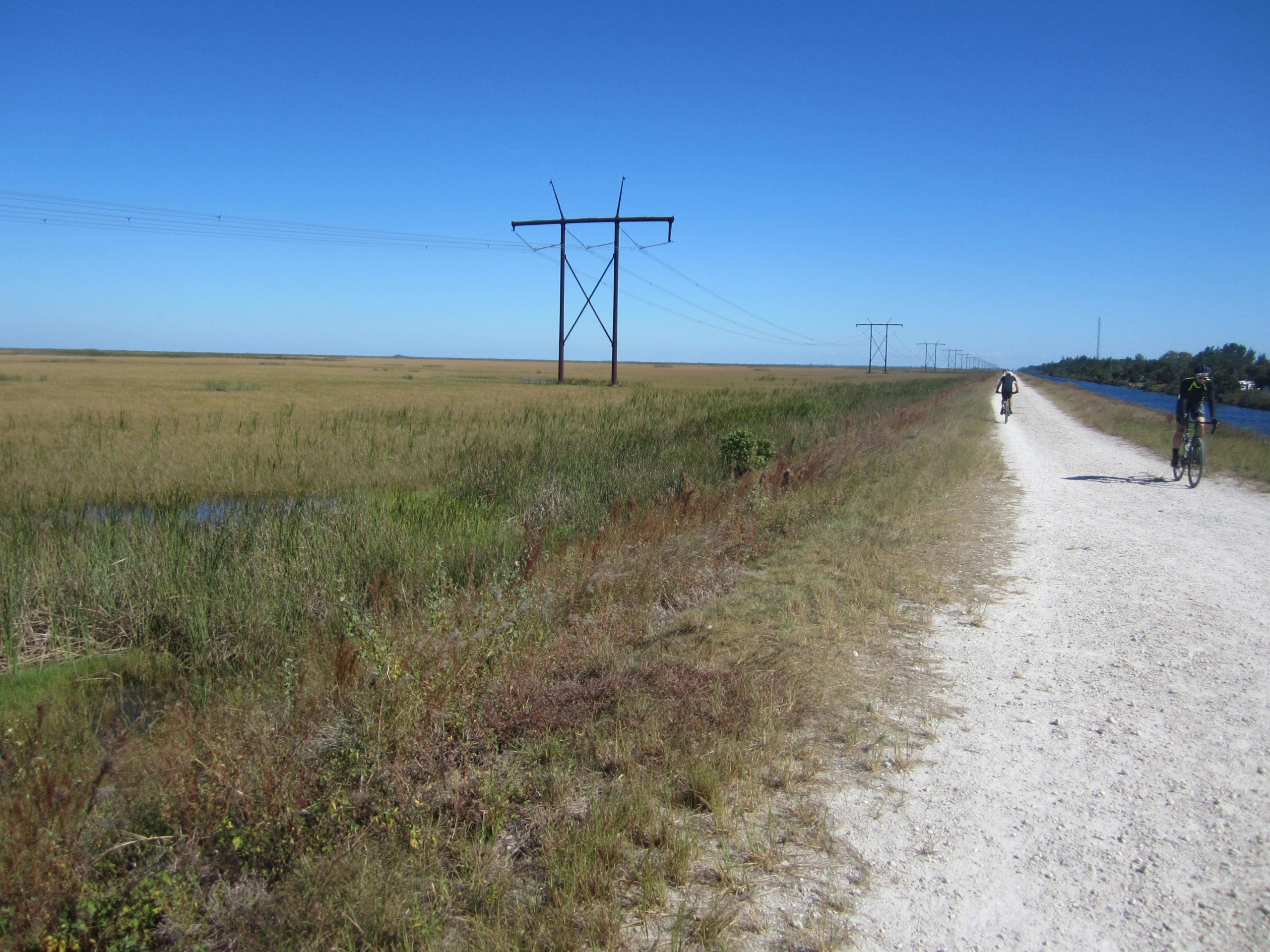 A wide, gravel path along a marshland area under a clear blue sky. Two cyclists ride on the trail, which is flanked by tall grasses and a power line running in the background. A narrow waterway is visible on the right side, reflecting the natural landscape. The Levee mountain bike trail.