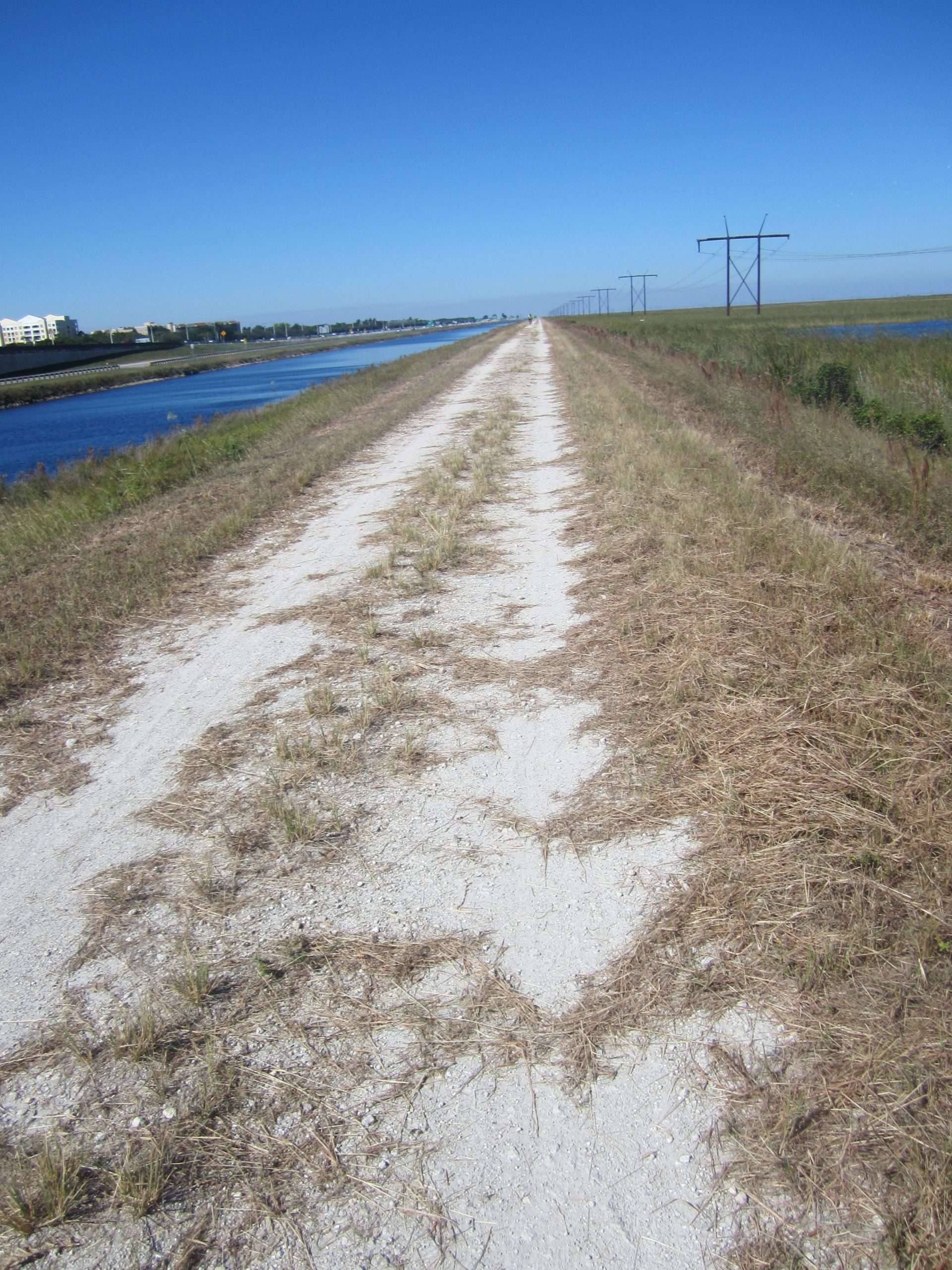 A long, unpaved dirt path lined with grass, running parallel to a canal on one side and power lines on the other, under a clear blue sky. The Levee mountain bike trail.