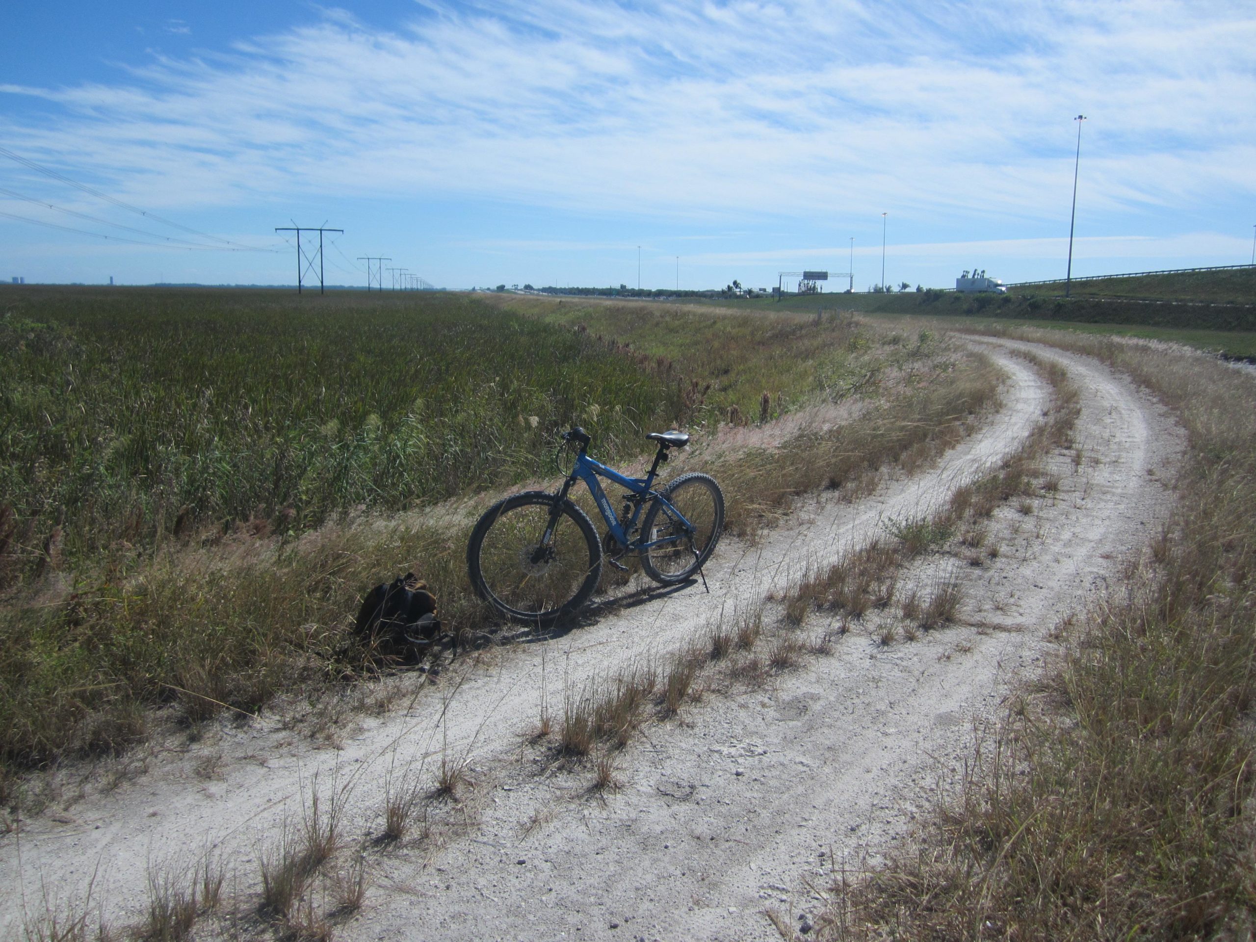 A blue mountain bike parked beside a dirt path, surrounded by tall grasses and a wide-open field under a clear blue sky. Power lines run in the distance, and a faint highway is visible on the right. A black backpack is resting on the ground near the bike. The Levee mountain bike trail.