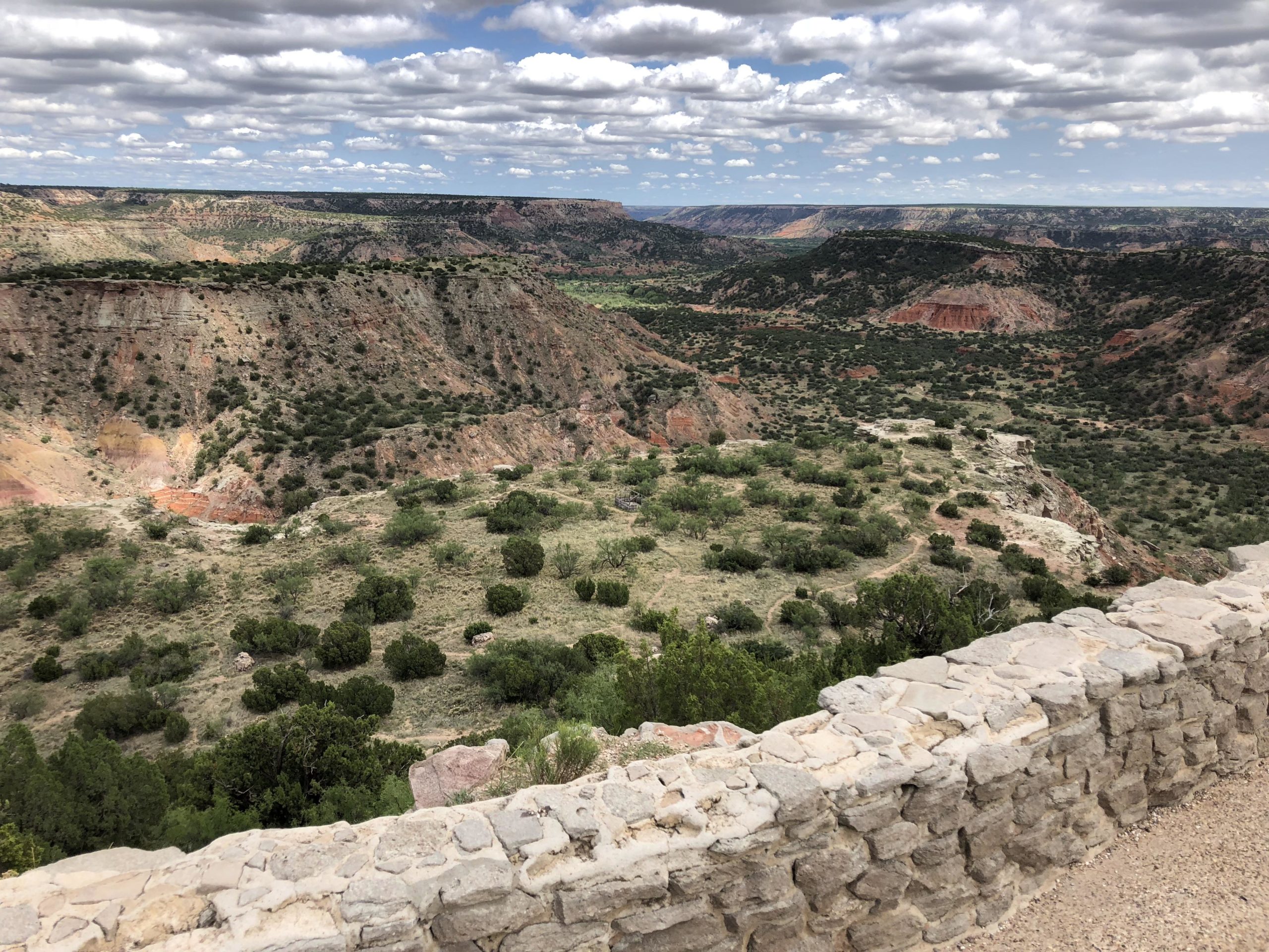 A scenic view of a canyon landscape featuring layered rock formations and lush greenery, under a partly cloudy sky. A stone wall is visible in the foreground, adding depth to the panoramic vista of rolling hills and canyons in the background. Palo Duro Canyon mountain bike trail.