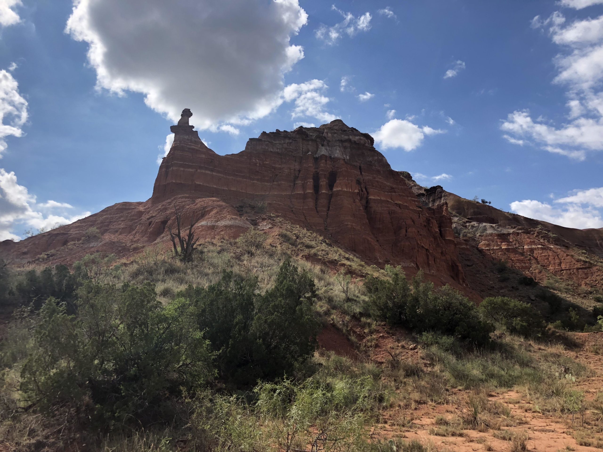 A rugged red rock formation towering under a blue sky dotted with fluffy clouds. The foreground features green vegetation, including shrubs and small bushes, while the rocky outcrop has a unique shape resembling a hoodoo, with a prominent cap at its peak. Palo Duro Canyon mountain bike trail.