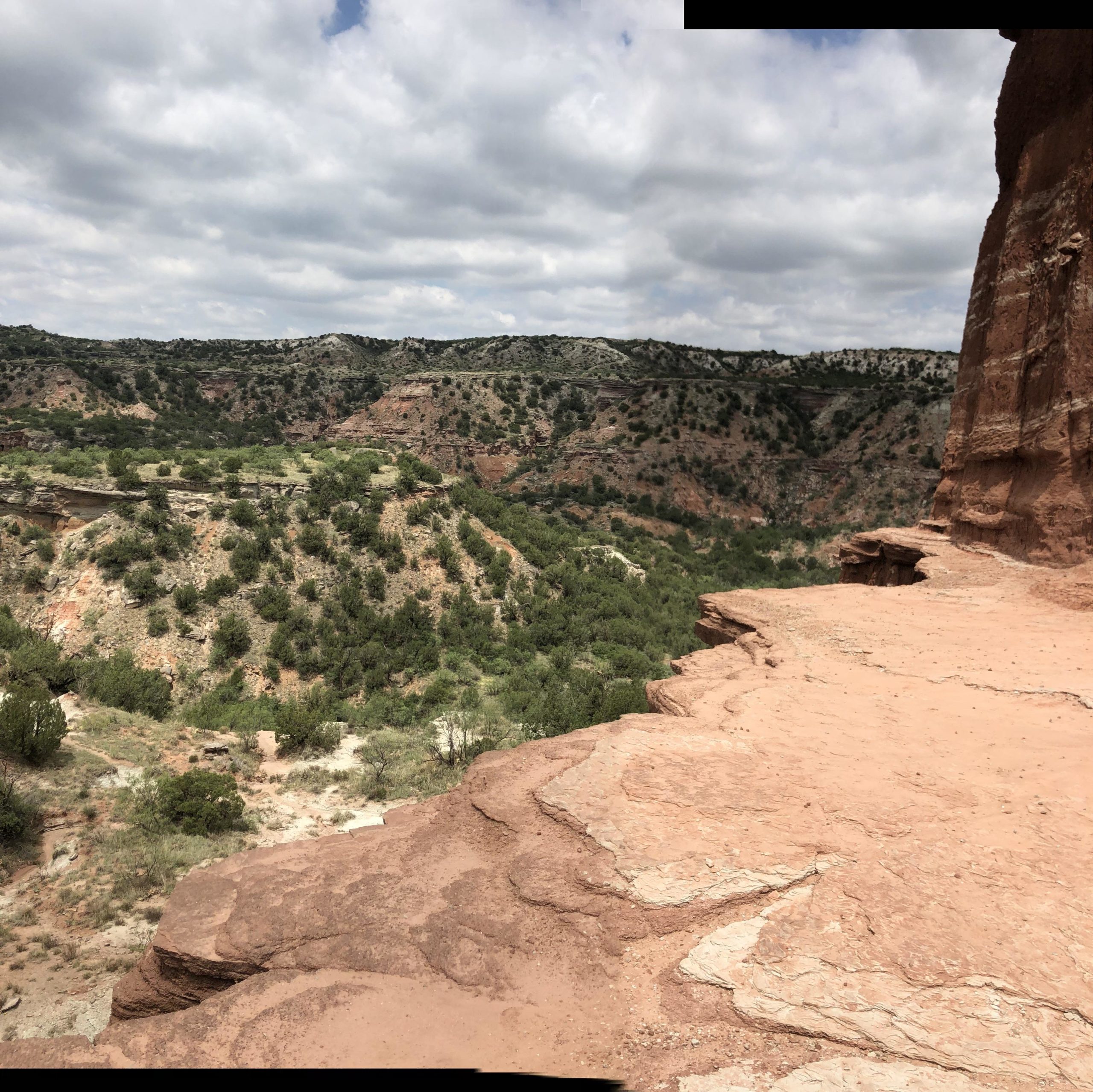 A panoramic view of rugged, layered rock formations and green vegetation in a canyon landscape under a partly cloudy sky. The foreground features rocky ledges with a smooth texture, while the background showcases rolling hills and cliffs. Palo Duro Canyon mountain bike trail.