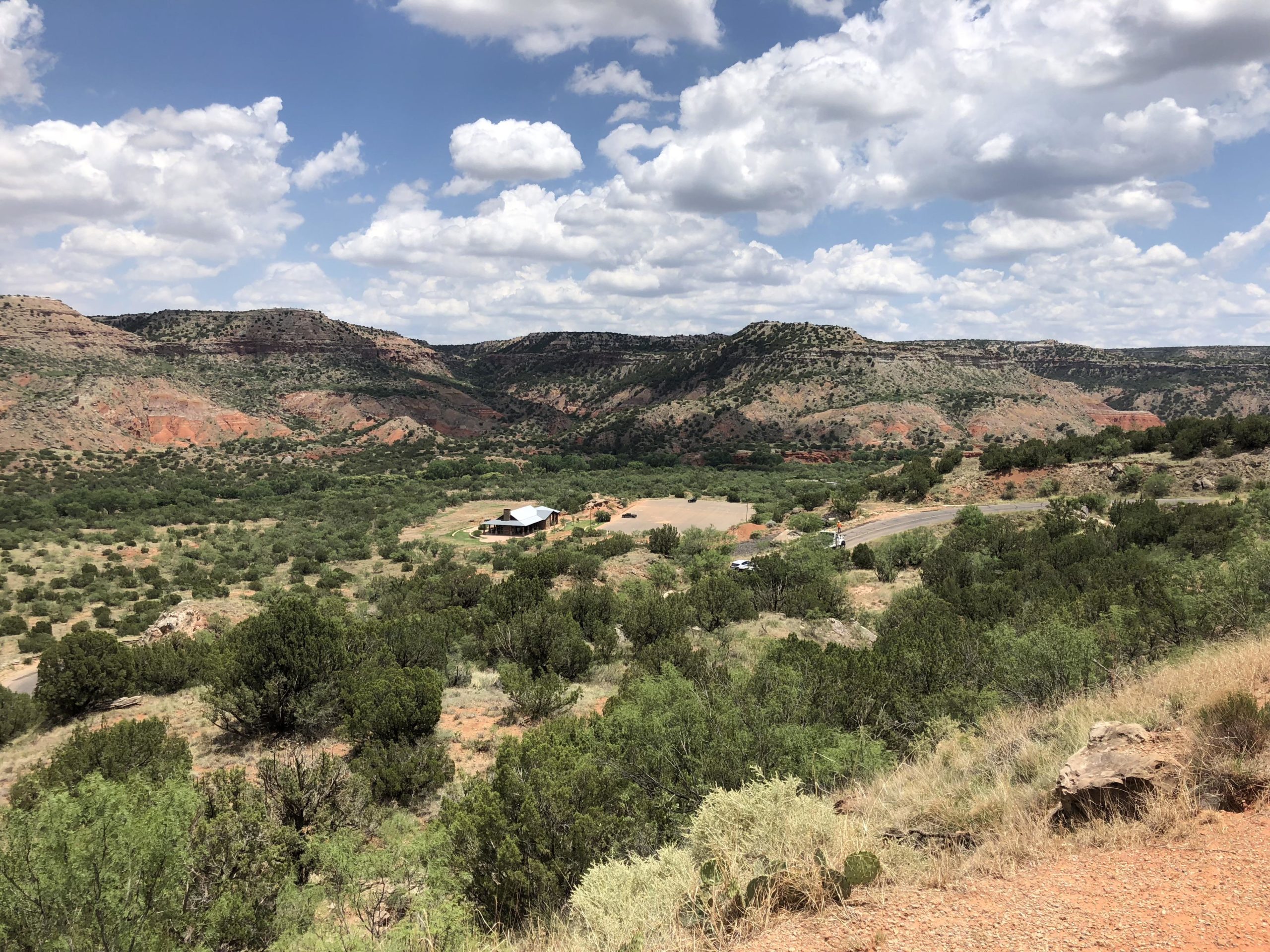 A panoramic view of a rugged landscape featuring green vegetation, rocky hills, and red cliffs under a partly cloudy sky. A small building is visible in the lower right corner, with a winding road in the distance. Palo Duro Canyon mountain bike trail.