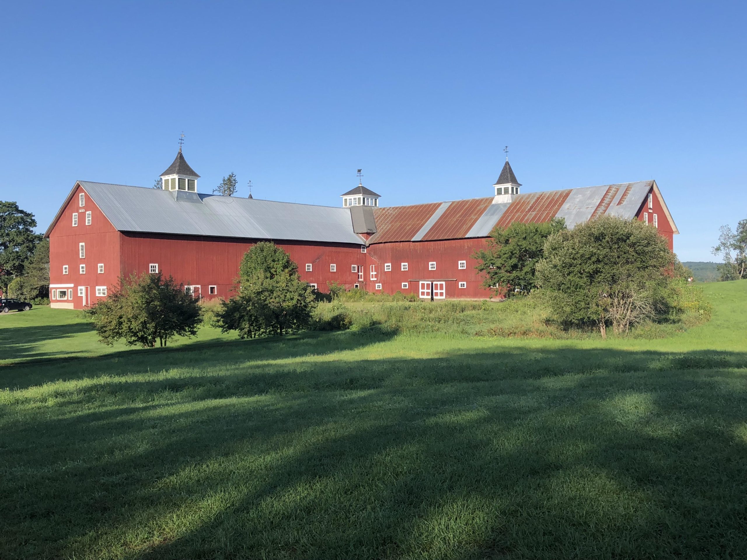 A large red barn with a metal roof and decorative cupolas, set against a clear blue sky. The barn is surrounded by lush green grass and small trees, emphasizing a rural landscape. Kingdom Trails mountain bike trail.