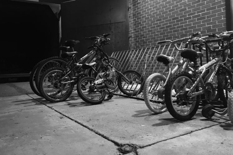 A collection of bicycles, including children's bikes and larger bikes, parked against a brick wall in a dimly lit area. The scene is captured in black and white, highlighting the textures of the bicycles and the concrete pavement.