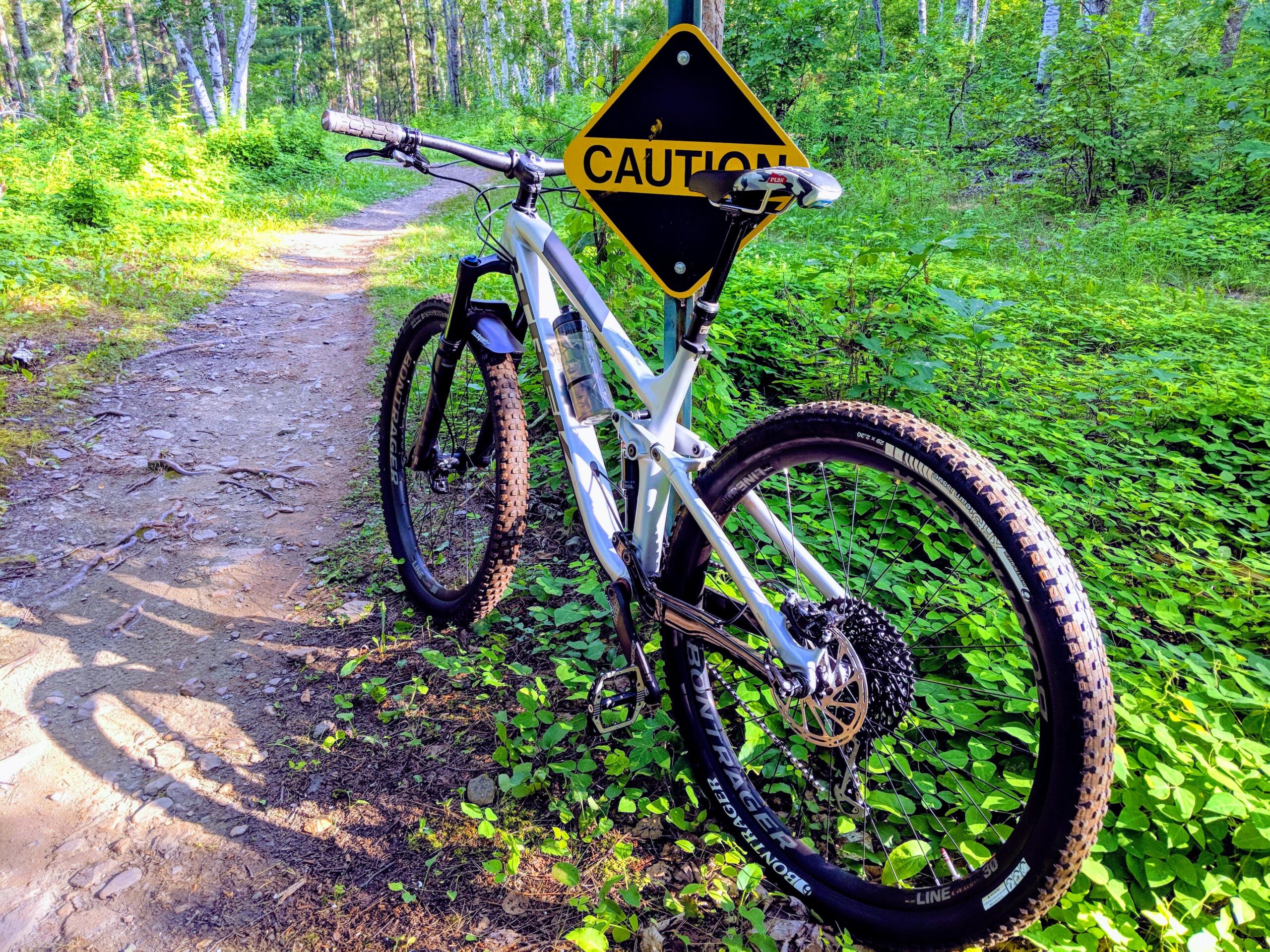 Trek Fuel EX 8: A mountain bike parked beside a trail sign that reads "CAUTION," surrounded by lush greenery and a dirt path. The bike is angled to show its tires and frame against the backdrop of a forested area.