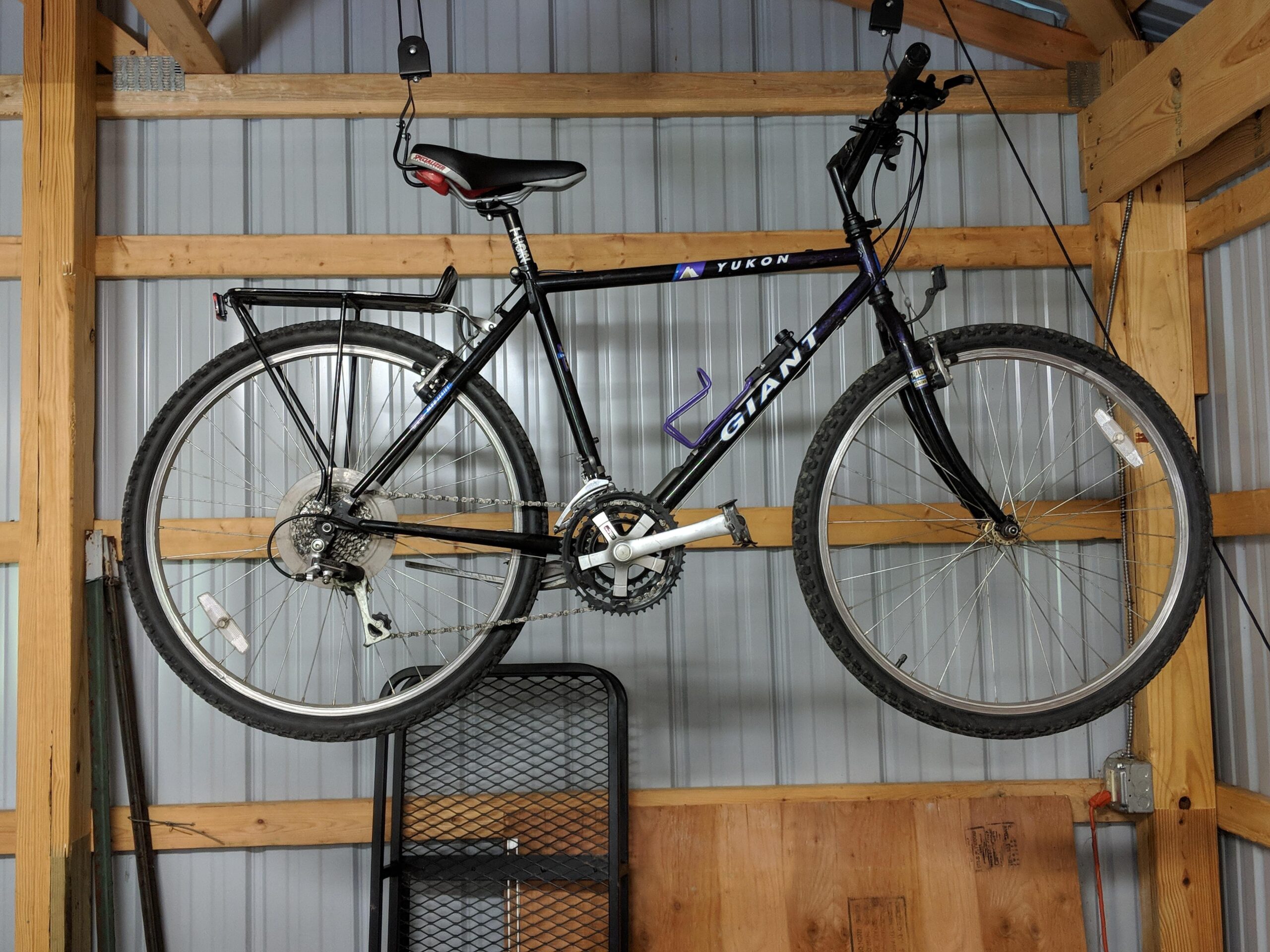 Giant Yukon: A black Giant Yukon mountain bike hanging from hooks in a wooden garage, with visible tires and gears. The background features metal walls and wooden beams, along with a black metal storage rack below the bike.