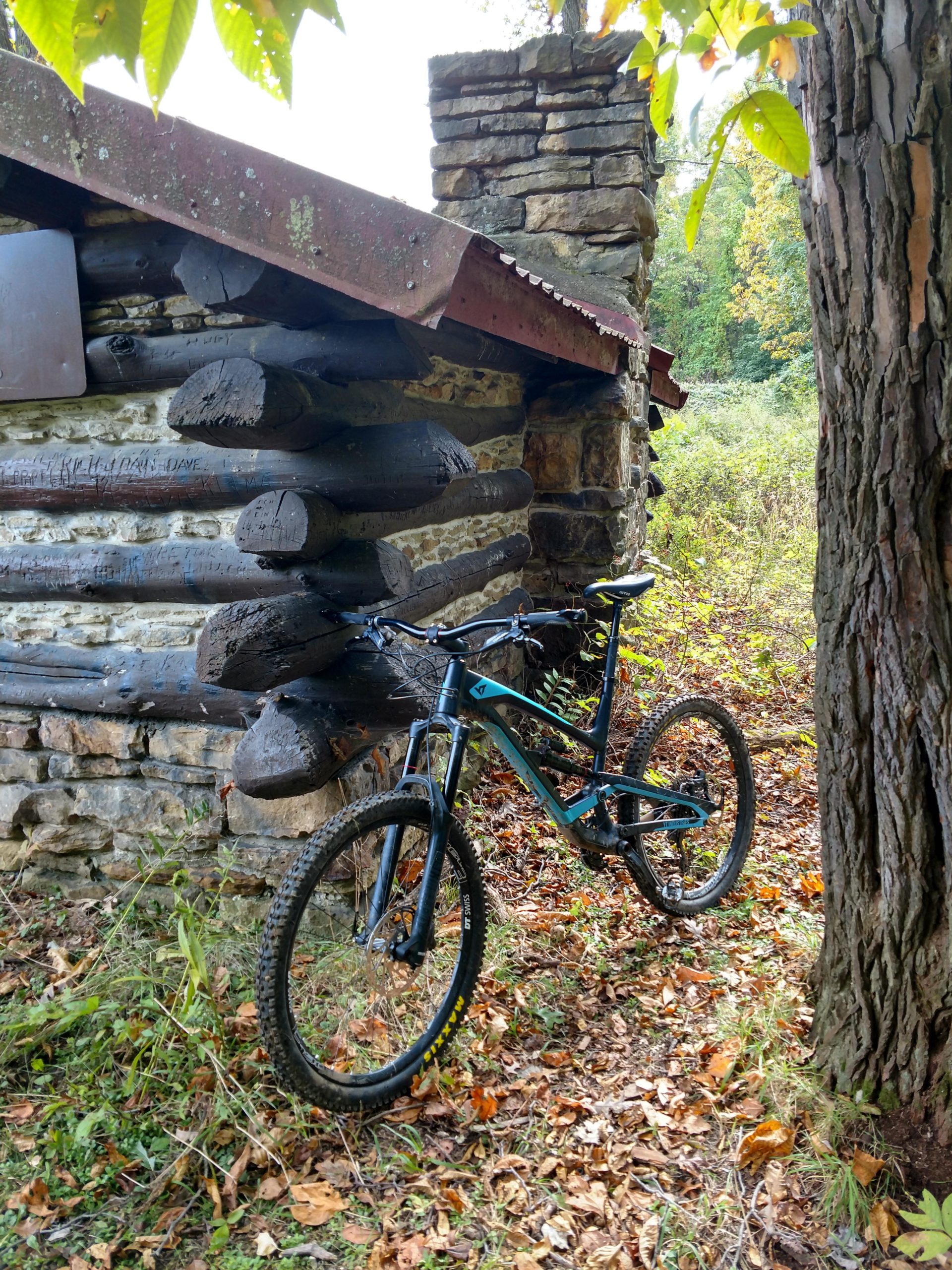 A mountain bike leaning against a rustic log cabin with a stone chimney, surrounded by autumn foliage and grasses. North Park mountain bike trail.