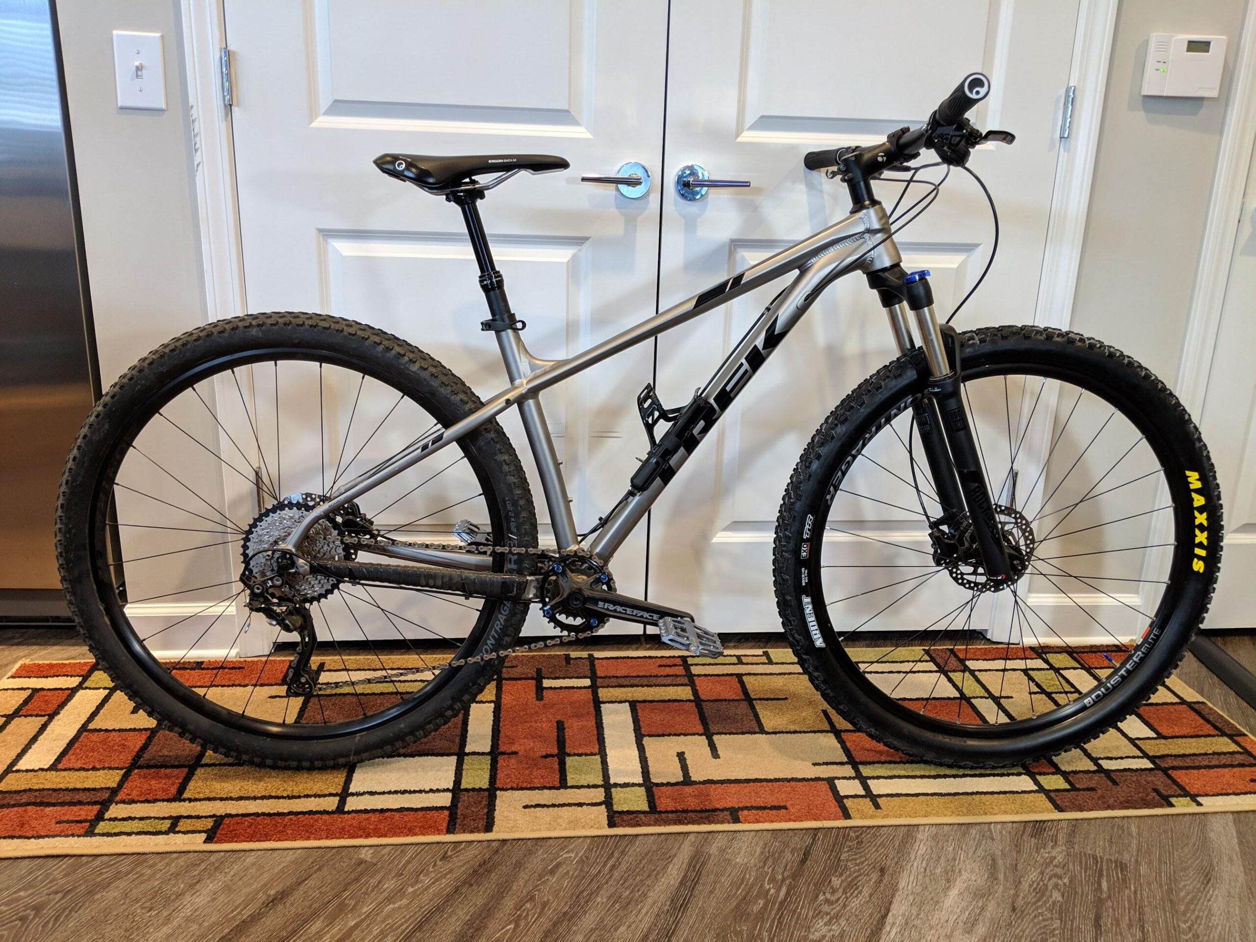Trek X-Caliber 8: A silver and black mountain bike is leaning against a wall with closed double doors in the background. The bike features large, knobby tires, a front suspension fork, and a modern frame design. It is positioned on a colorful geometric-patterned rug, and a stainless steel refrigerator is visible to the left.